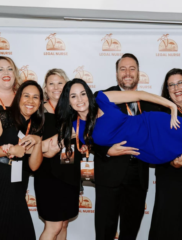 Group of six people, including a man holding a woman (Janice "Jan Marie" Dolnick) horizontally, smiling in front of a backdrop with 'Legal Nurse' logo.