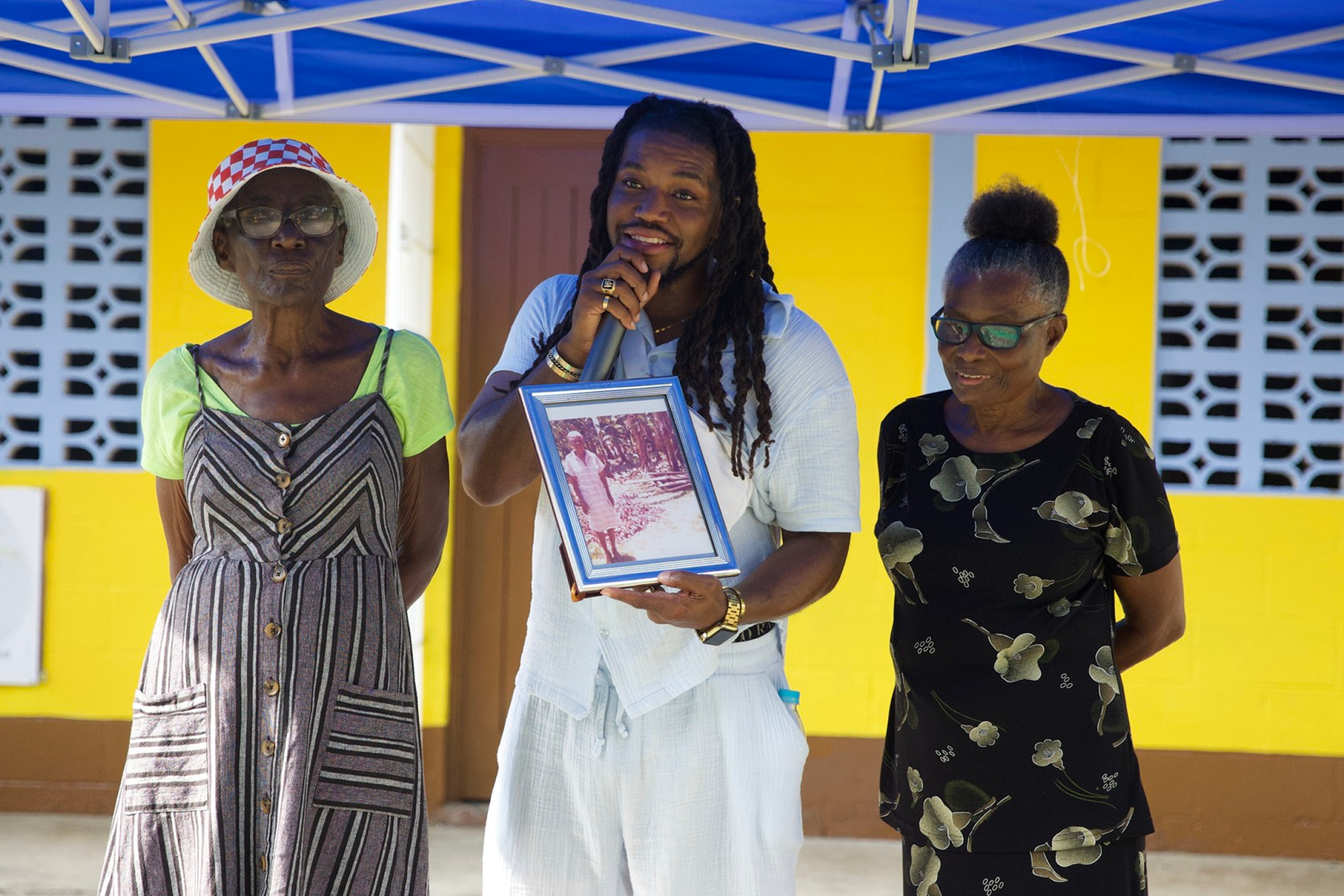 Three women standing together outdoors under a blue canopy, with a yellow wall behind them. The woman in the middle is holding a framed photo and speaking into a microphone. The women are dressed in casual summer clothes, and the scene appears to be part of a community event or gathering.