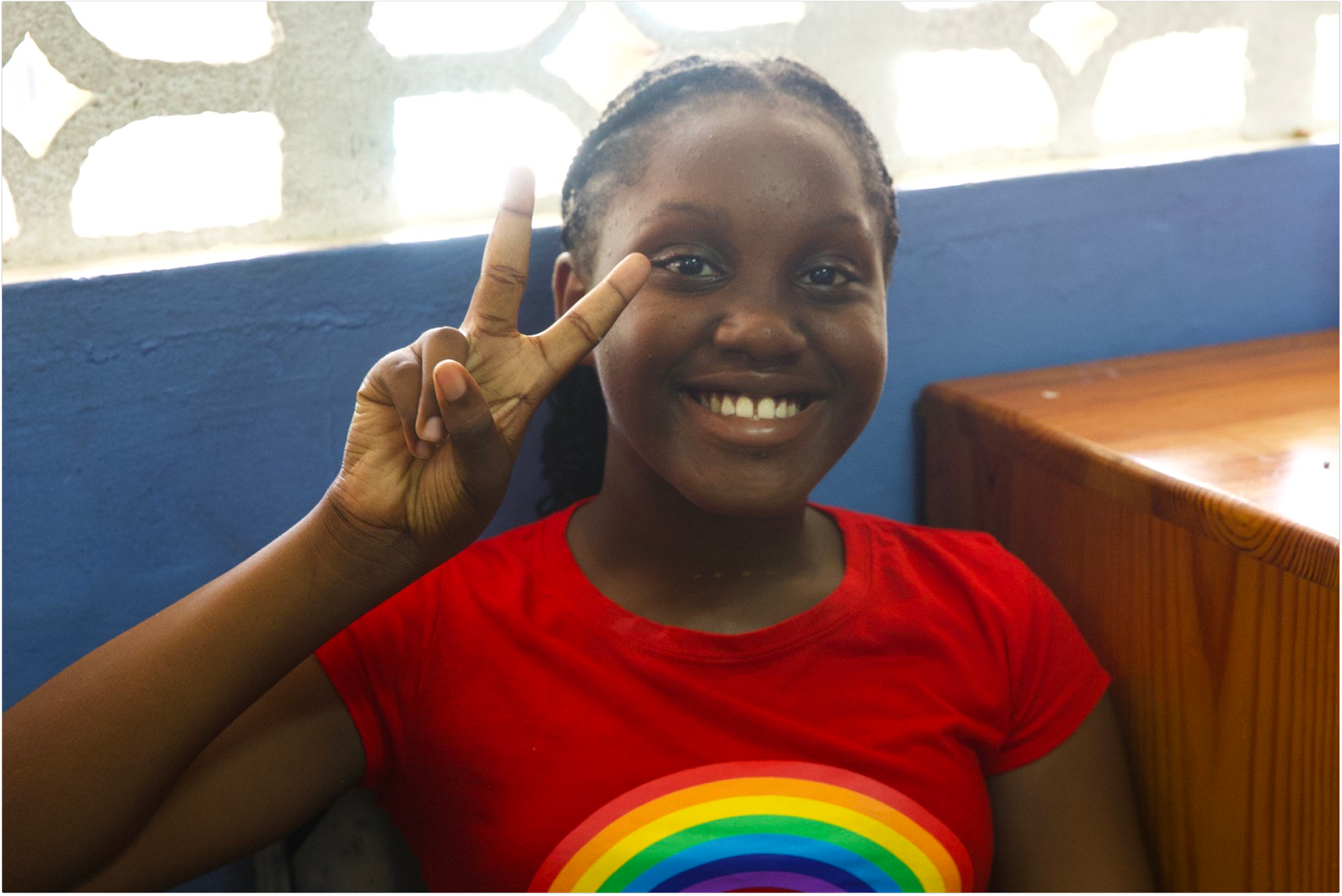 A young girl smiling and making a peace sign near her eye, wearing a red shirt with a rainbow graphic on it.