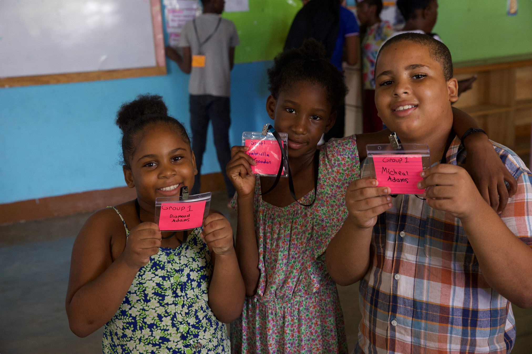 Three children smiling and holding up name tags at school, with other children and a teacher in the background.