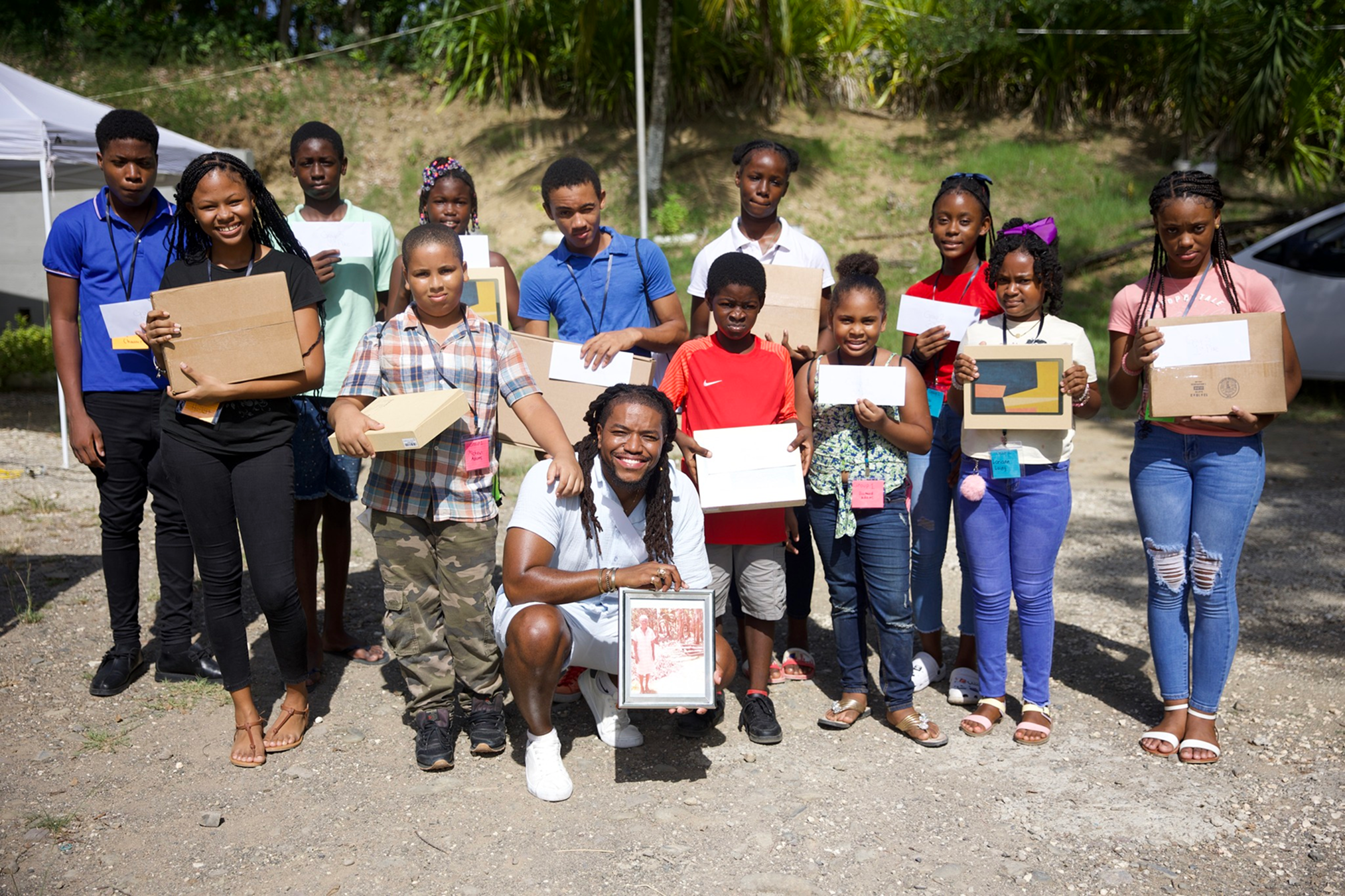 A group of children and an adult posing outdoors on a sunny day, holding various boxes and papers, with some smiling and others with neutral expressions, in front of a hillside with trees and a white tent.