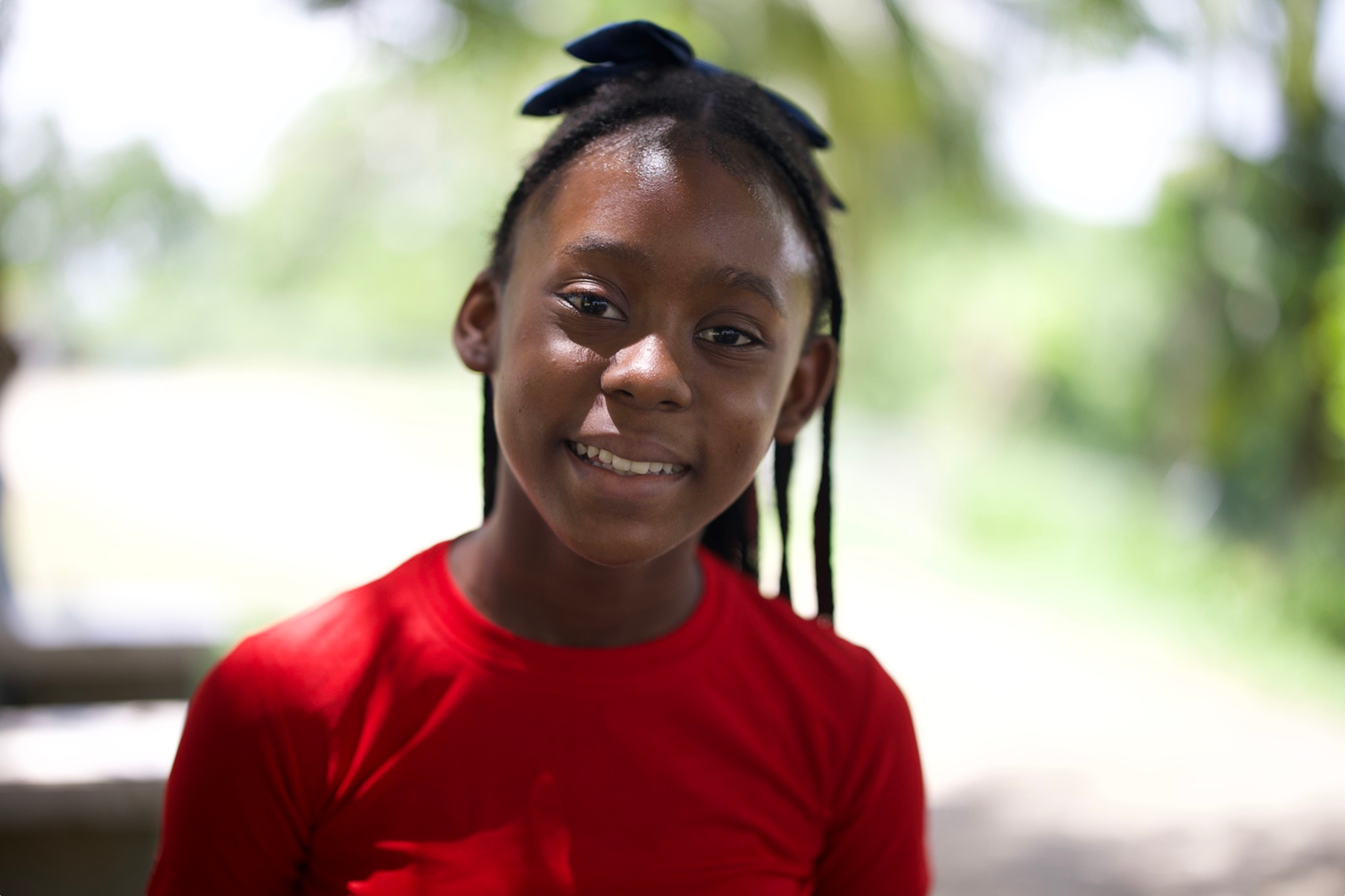 A young girl with braided hair wearing a red shirt, smiling outdoors with blurred green foliage in the background.