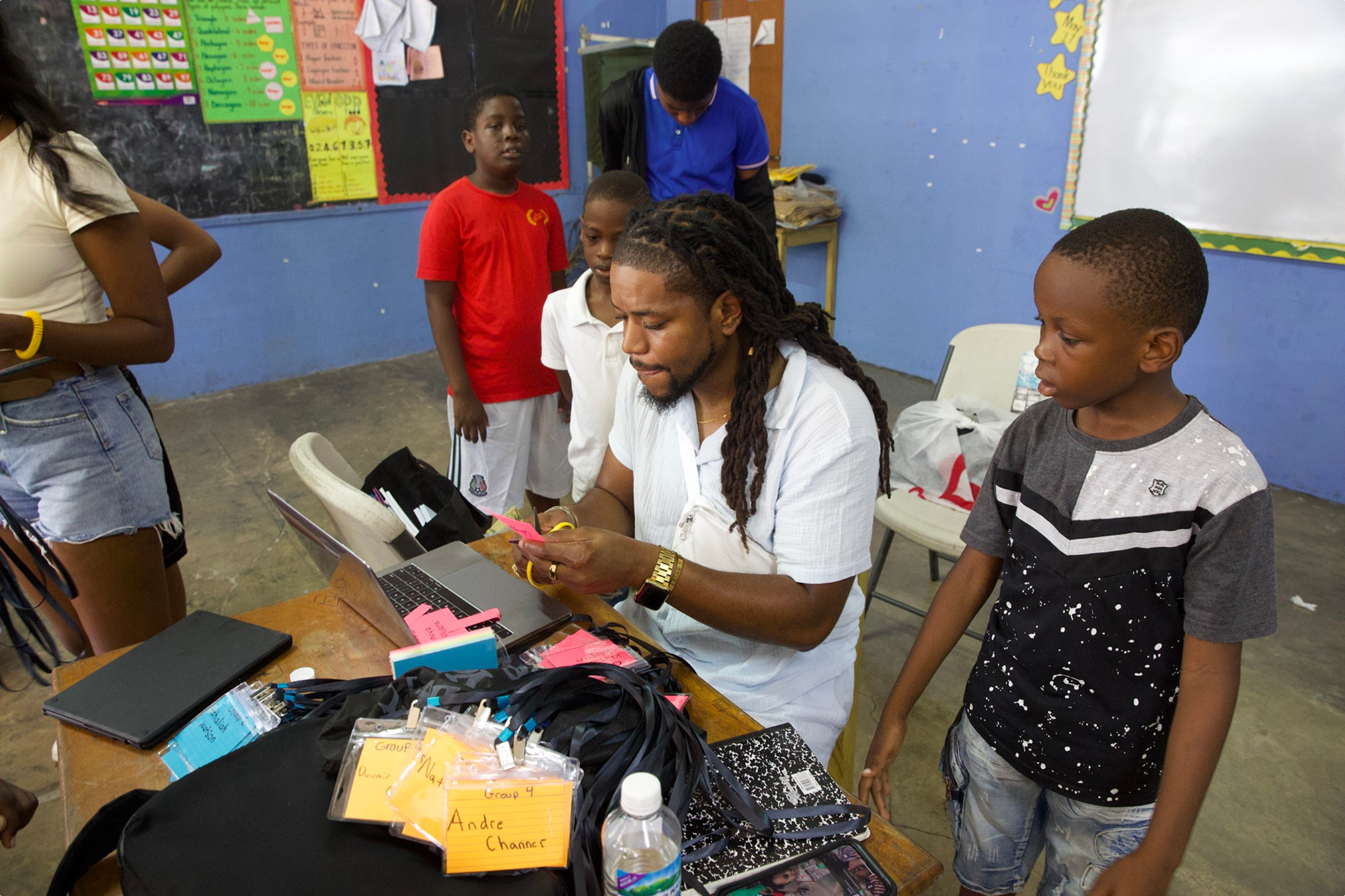 A classroom scene with a man sitting at a desk surrounded by children. The man is looking at a smartphone, and the children are standing around him, observing. The desk has various school supplies, including notebooks, name tags, and a water bottle. The classroom has colorful educational posters on the walls and a blue background.