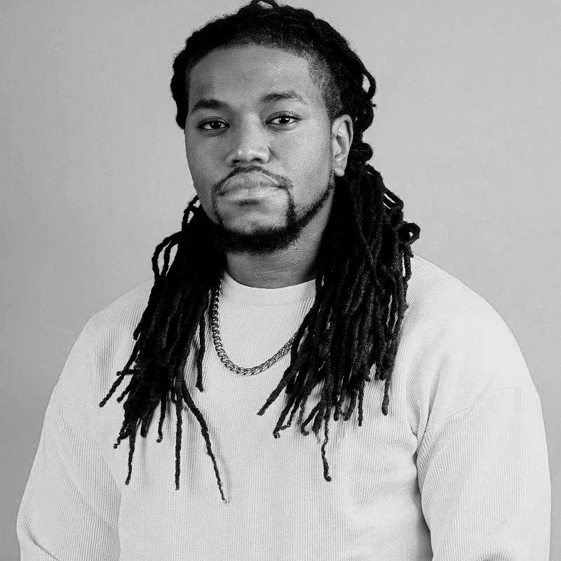 A man with long dreadlocks, a beard, and a mustache, wearing a white sweater and a chain necklace, posing against a plain background.