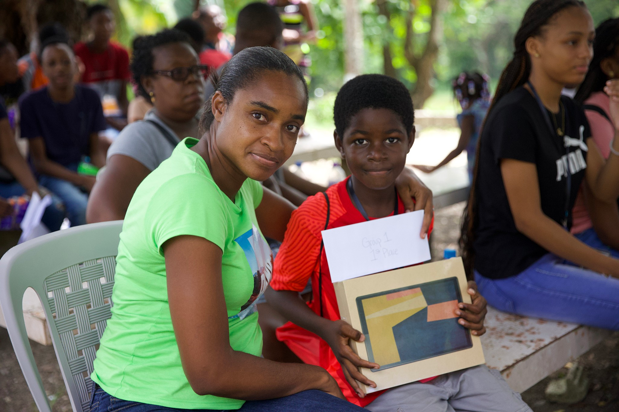 A woman and a boy sitting outdoors among other children, with the woman embracing the boy. The boy holds a tablet displaying colorful blocks and a white paper that reads 'Group 1 1st Place.' The scene takes place in a lush, green, shaded area during the daytime, and the children are dressed casually.