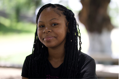 A young girl with long braided hair smiling outdoors on a sunny day.
