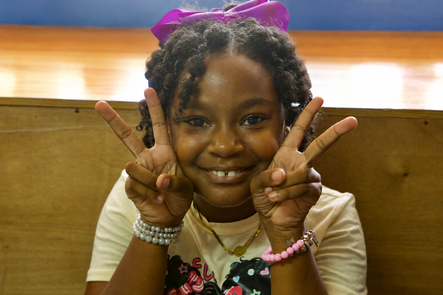 A young girl with curly hair tied with a purple bow, making peace signs with her fingers, smiling, wearing jewelry, seated against a wooden background.