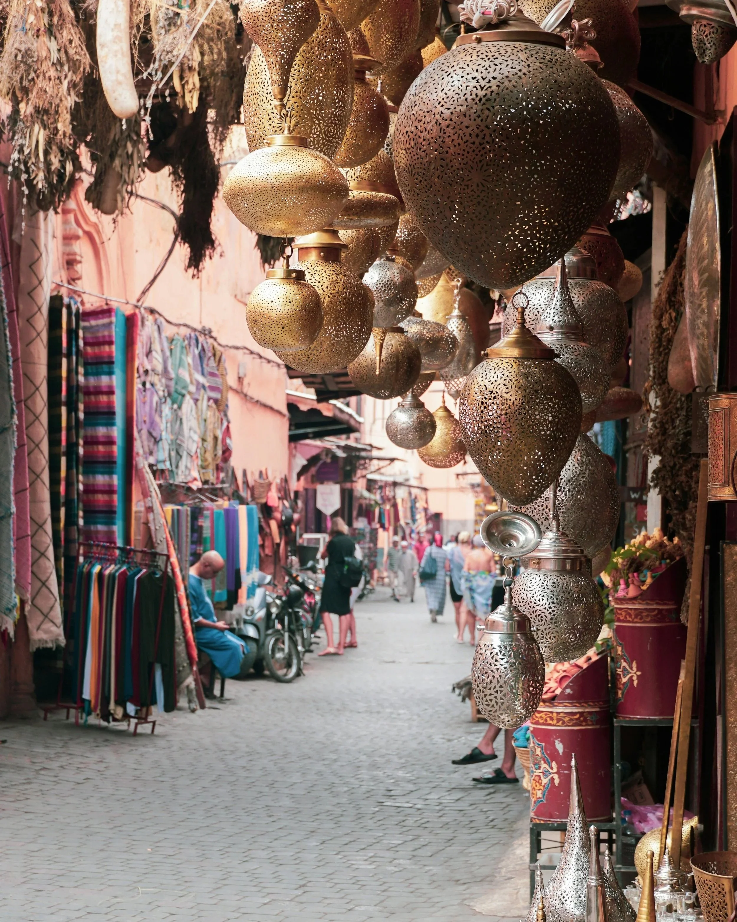 Market street with hanging decorative metal lamps and various textiles on display, and people walking and browsing shops.