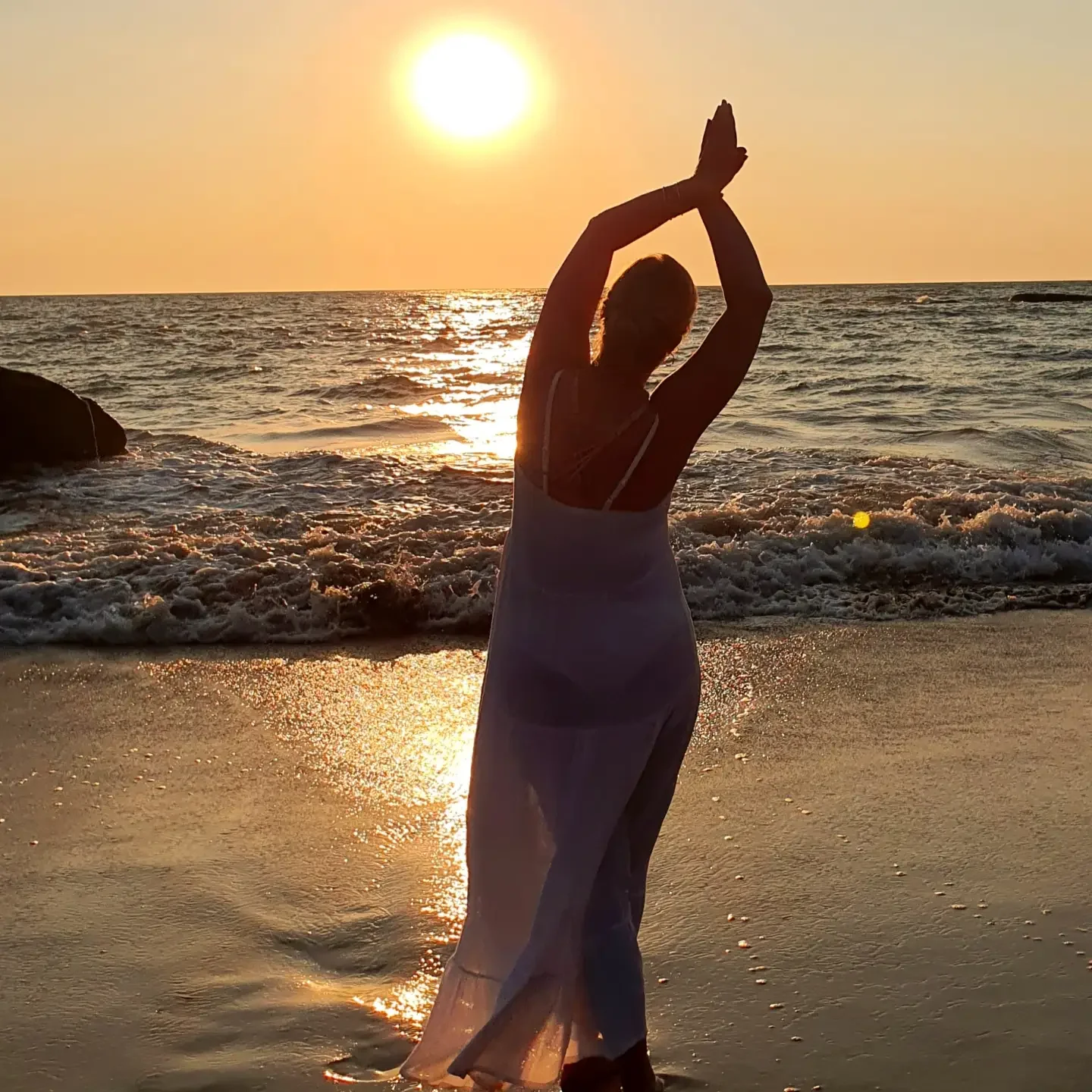 A woman practicing yoga on the beach at sunset, with her arms raised above her head in a yoga pose, facing the ocean.