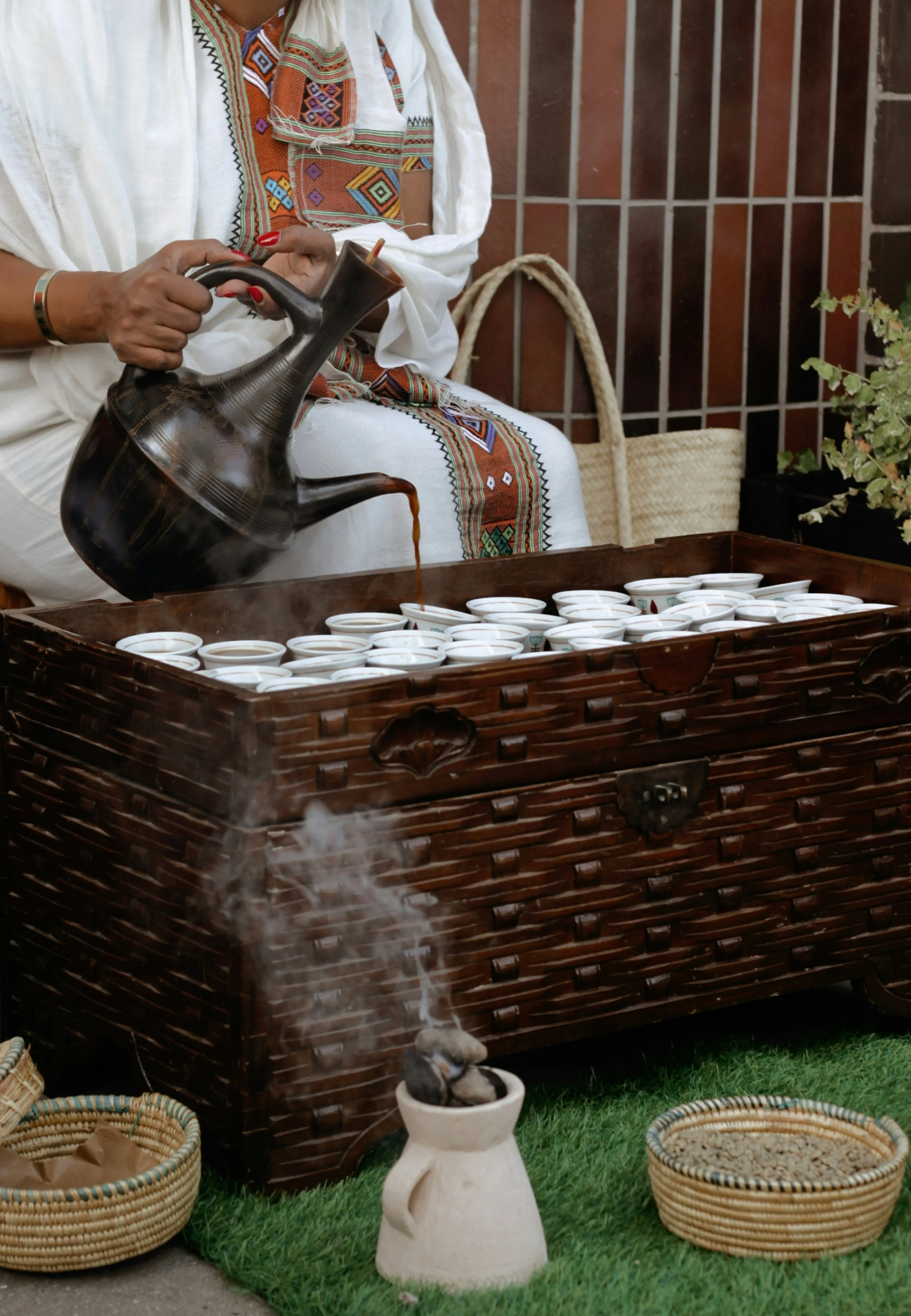 A person in traditional clothing sitting on a wooden chair and pouring a dark beverage from a ceramic pitcher into small cups on a tray. The scene is set outdoors on grass, with baskets containing various items nearby and decorative plants and a brick wall in the background.