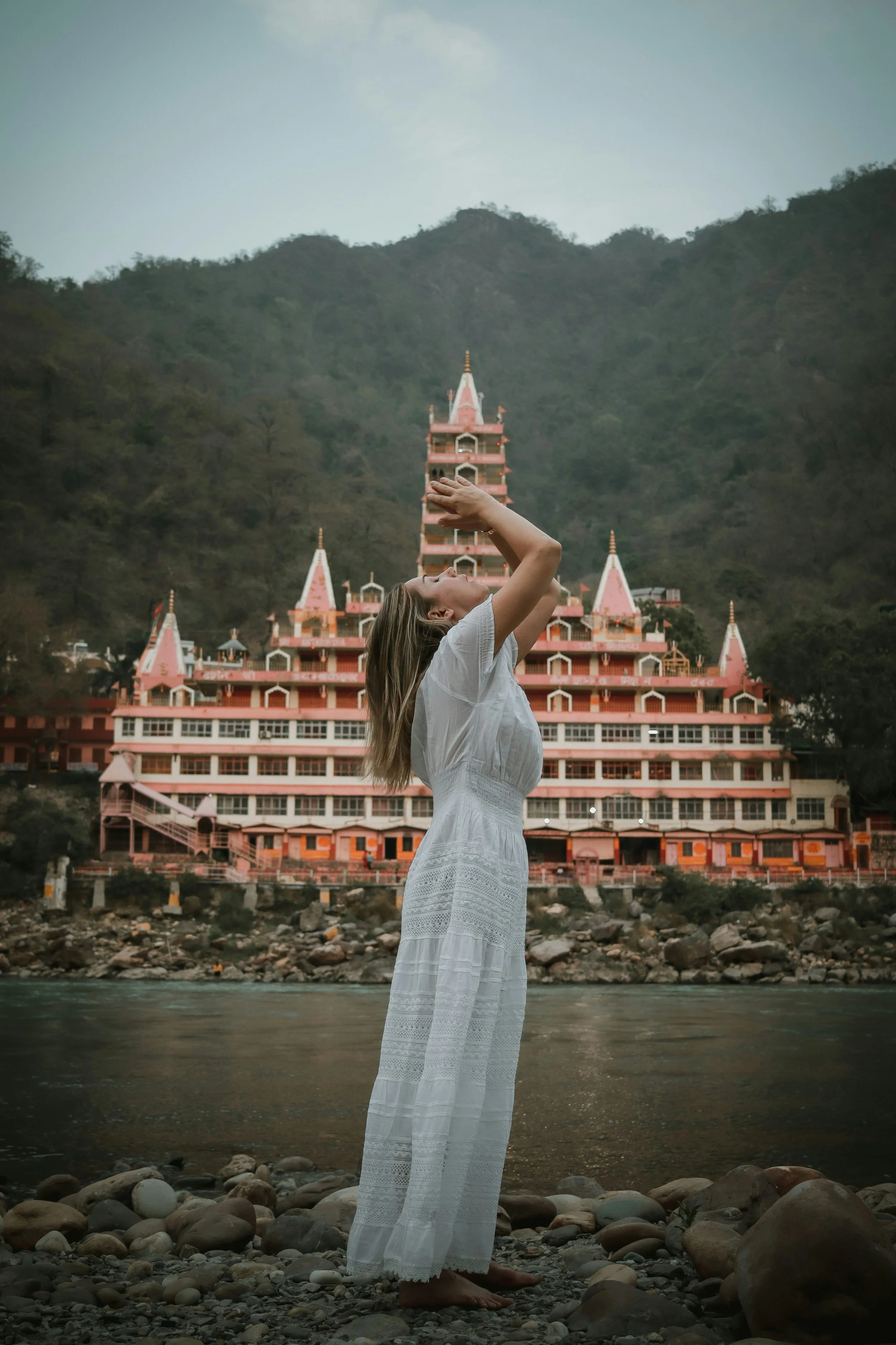 A woman in a white dress standing barefoot on rocky riverbank, holding her hands above her head, with a pink temple in the background and mountains behind.