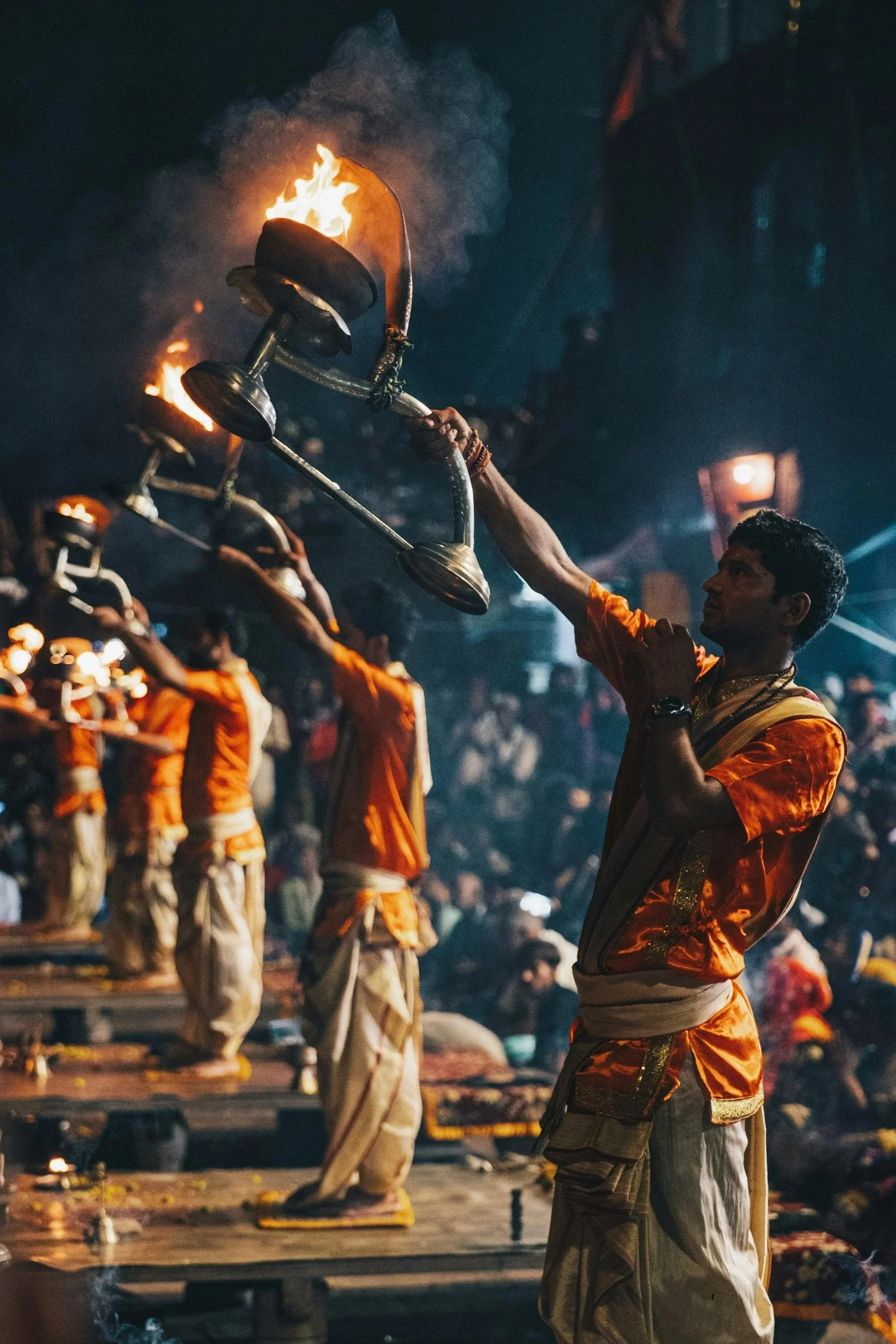 A group of men dressed in traditional Indian attire participating in a religious or cultural festival at night, holding a lit brass lamp with flames.