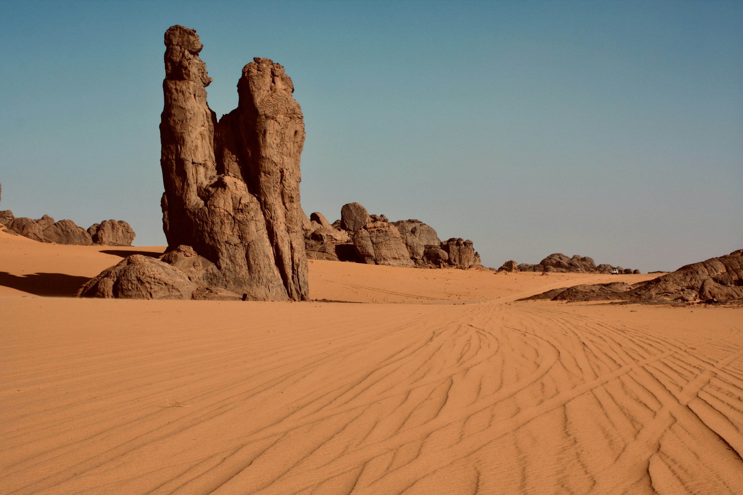 Desert landscape with large rock formations, sand dunes, and a small vehicle in the distance under a clear blue sky.