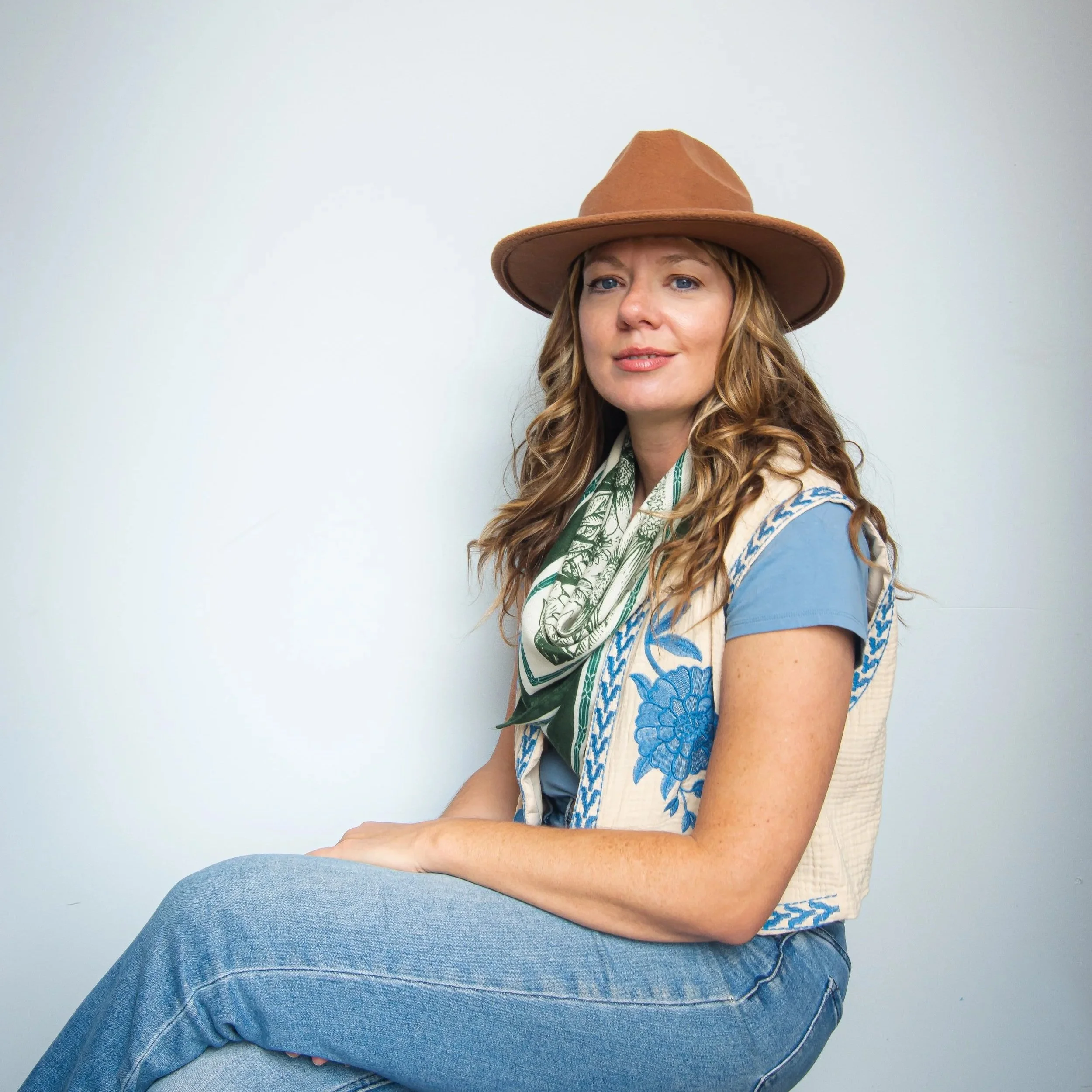 A woman with long, wavy blonde hair wearing a brown wide-brimmed hat, a blue patterned shirt, and blue jeans, sitting sideways with her arms crossed, against a plain white wall.