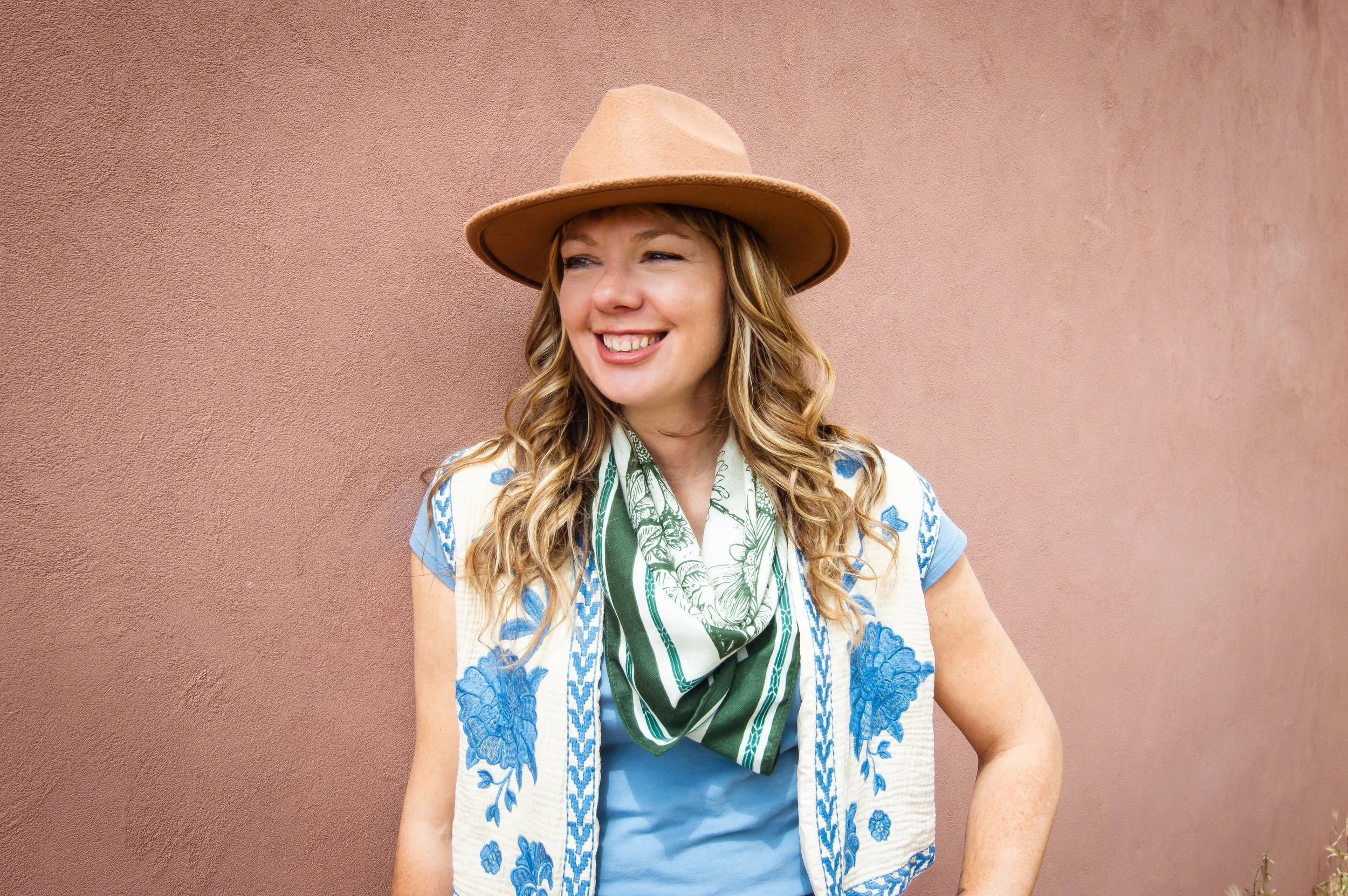 A woman smiling and looking to her left, wearing a large tan hat, a light blue top, a white vest with blue embroidered floral patterns, and a green and white patterned scarf, standing against a pinkish wall.
