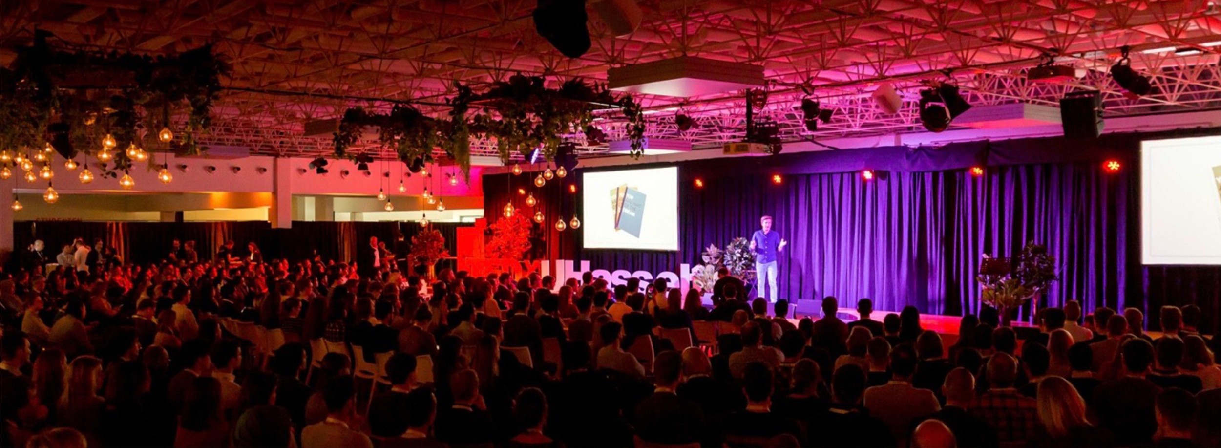 A large conference or concert hall filled with seated audience watching a speaker on stage. The stage has a large screen displaying a presentation and is decorated with plants. The hall has a high ceiling with hanging lights and pink and purple lighting.