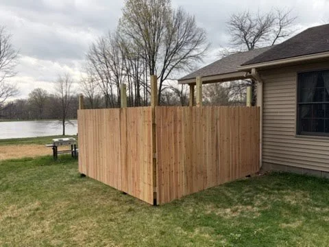 Newly installed wooden privacy fence surrounding the backyard corner of a house, with a lawn and trees in the background.
