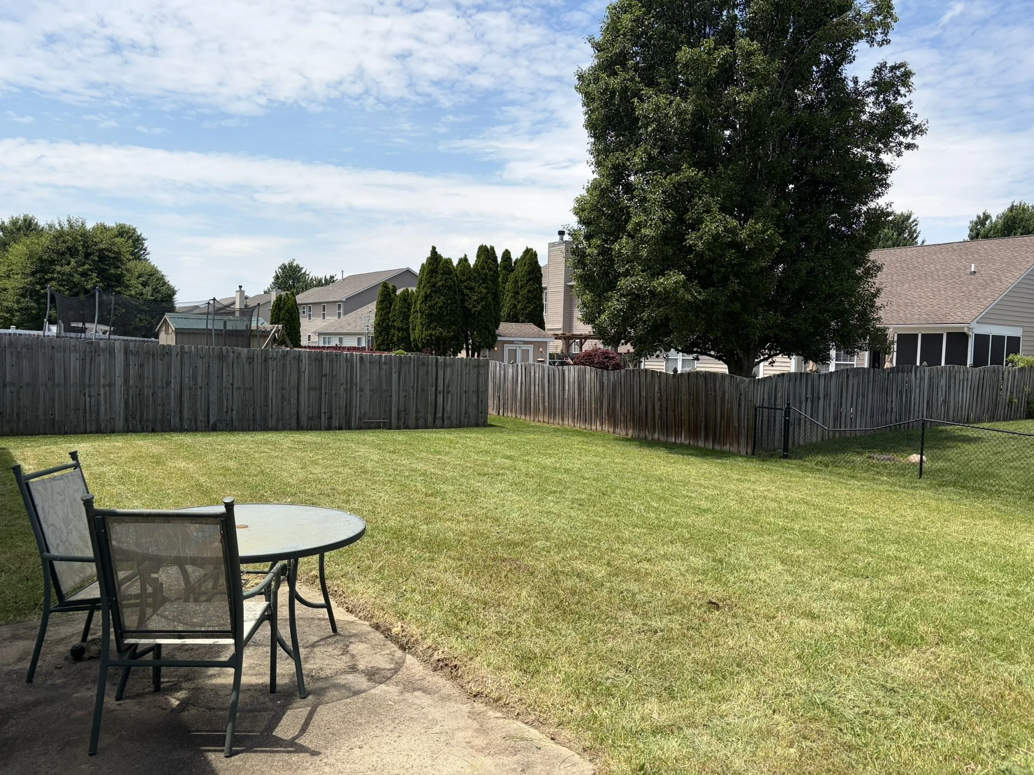 Backyard with grass, patio furniture, a wooden fence, trees, houses, and a partly cloudy sky.