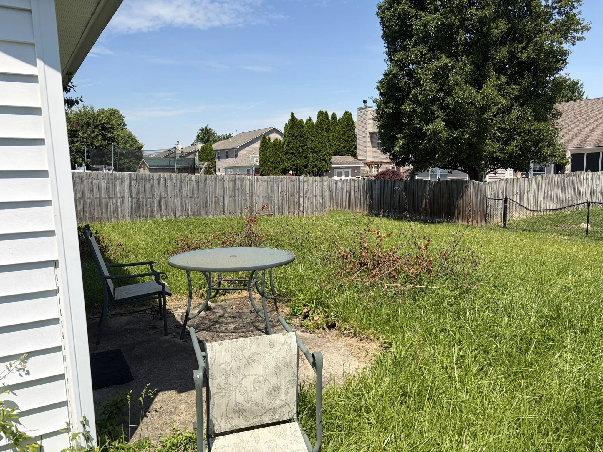 Backyard scene with a patio area, metal table and chairs, grassy lawn, wooden fence, large tree, and neighboring houses with clear blue sky.