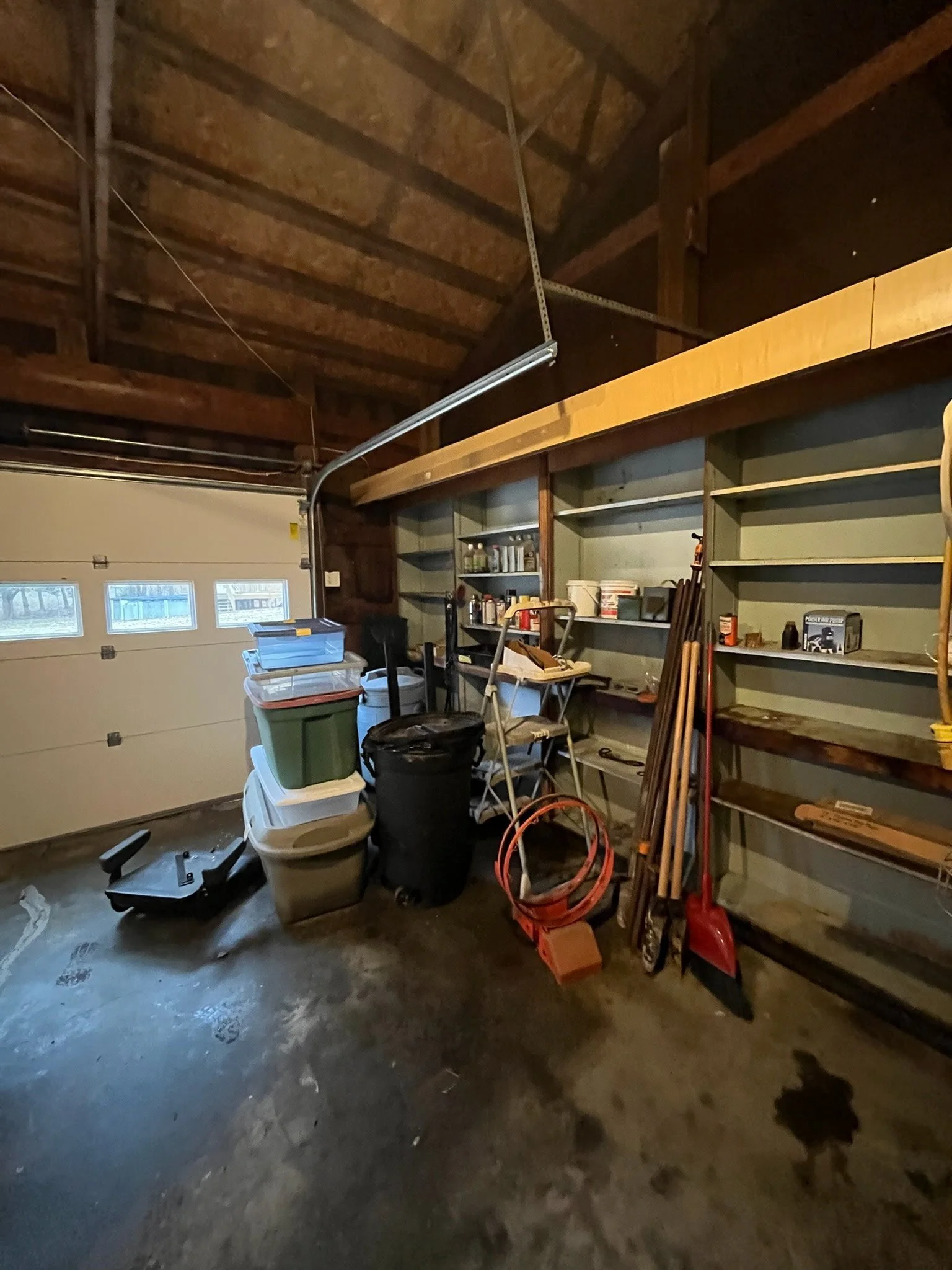 A cluttered garage with shelves filled with various household items, gardening tools, and storage bins, with a concrete floor and a partially visible garage door.