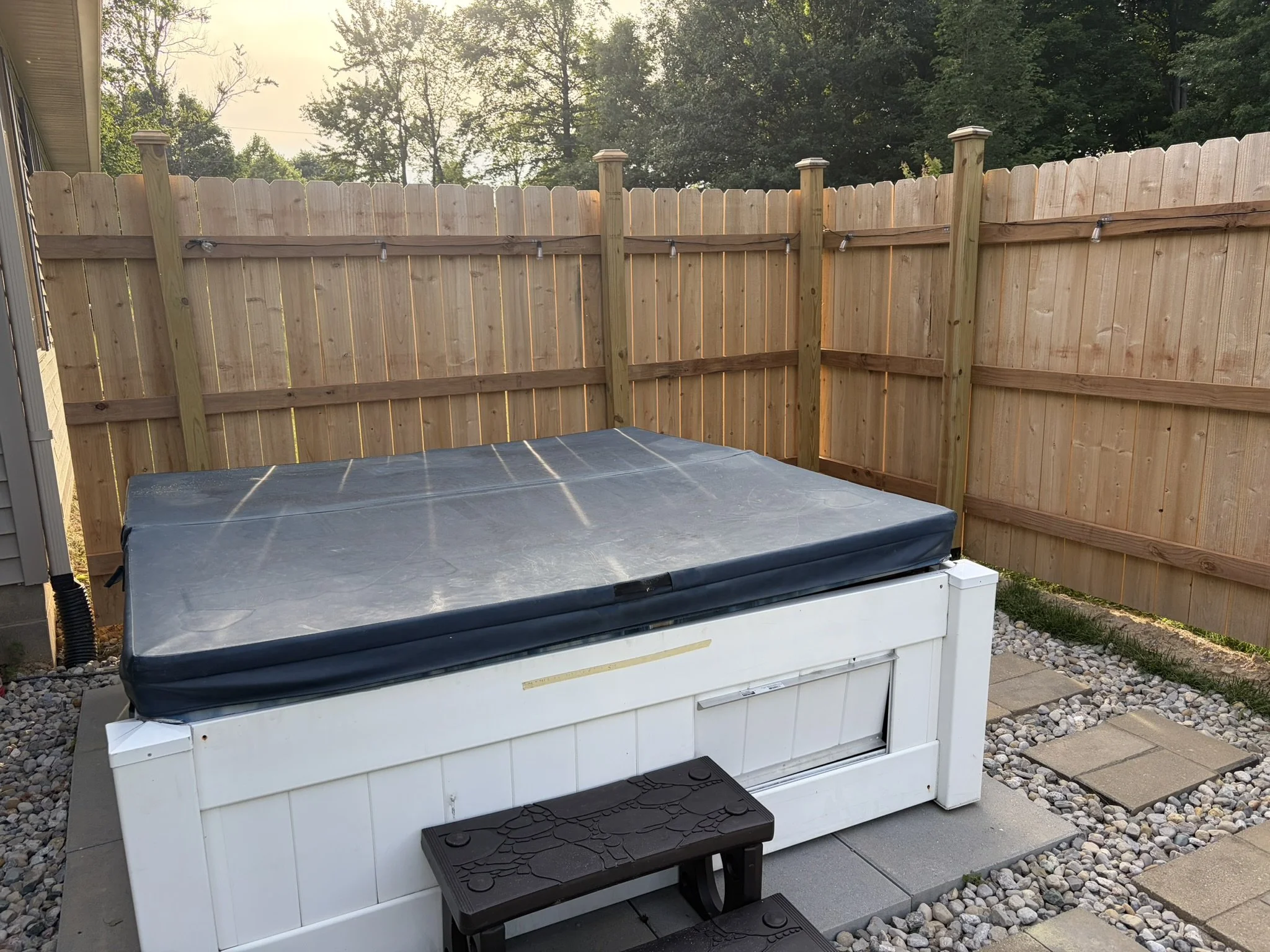 A wooden privacy fence in a backyard, with some string lights attached, surrounding a hot tub and a storage box on a concrete patio, with trees and blue sky in the background.