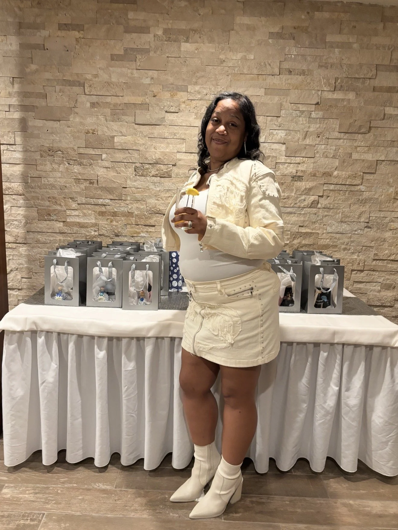 Woman in cream-colored jacket and shorts holding a drink, standing in front of a table with gift bags, in a room with beige brick wall.