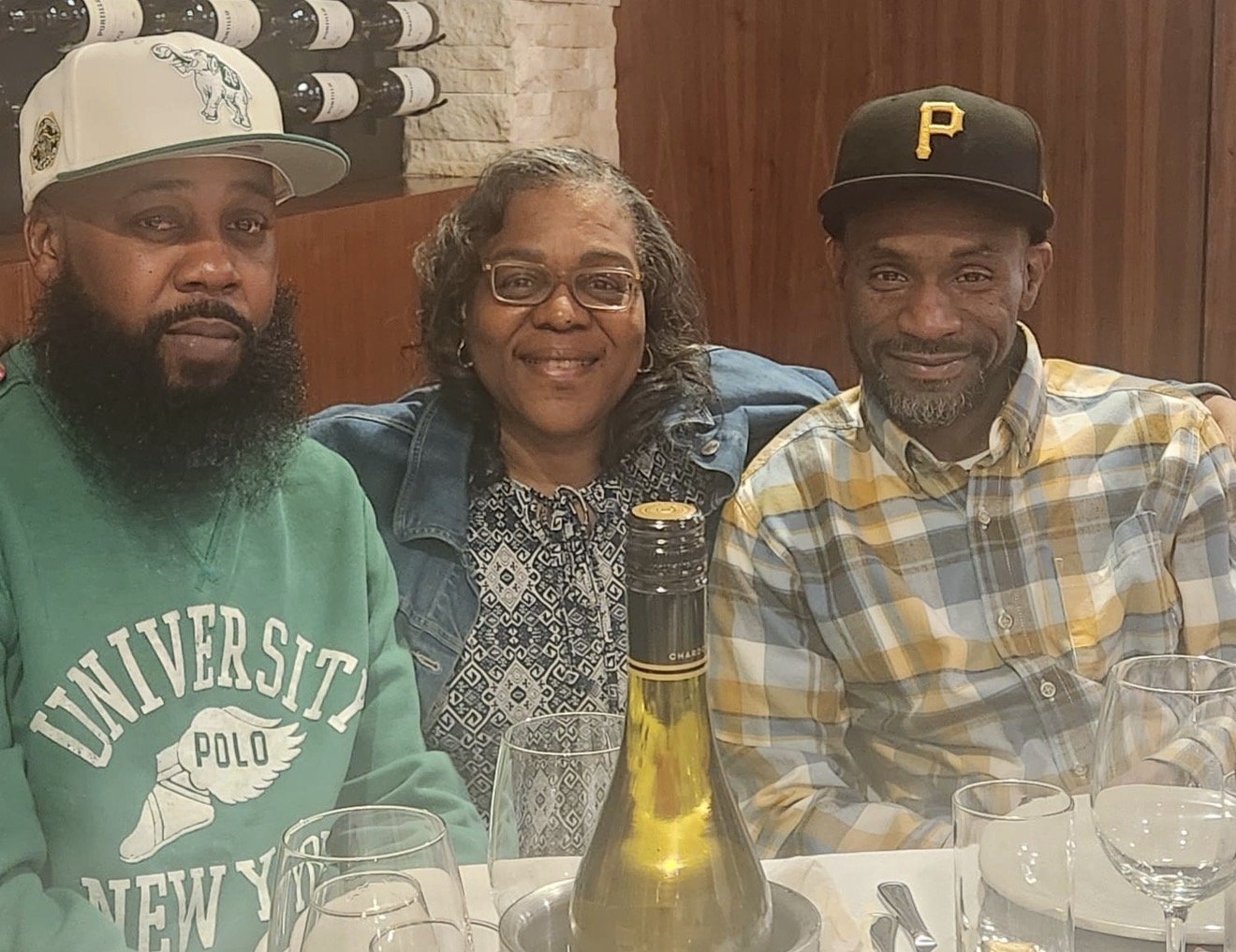 Three people sitting at a restaurant table with wine glasses and a bottle, smiling at the camera.