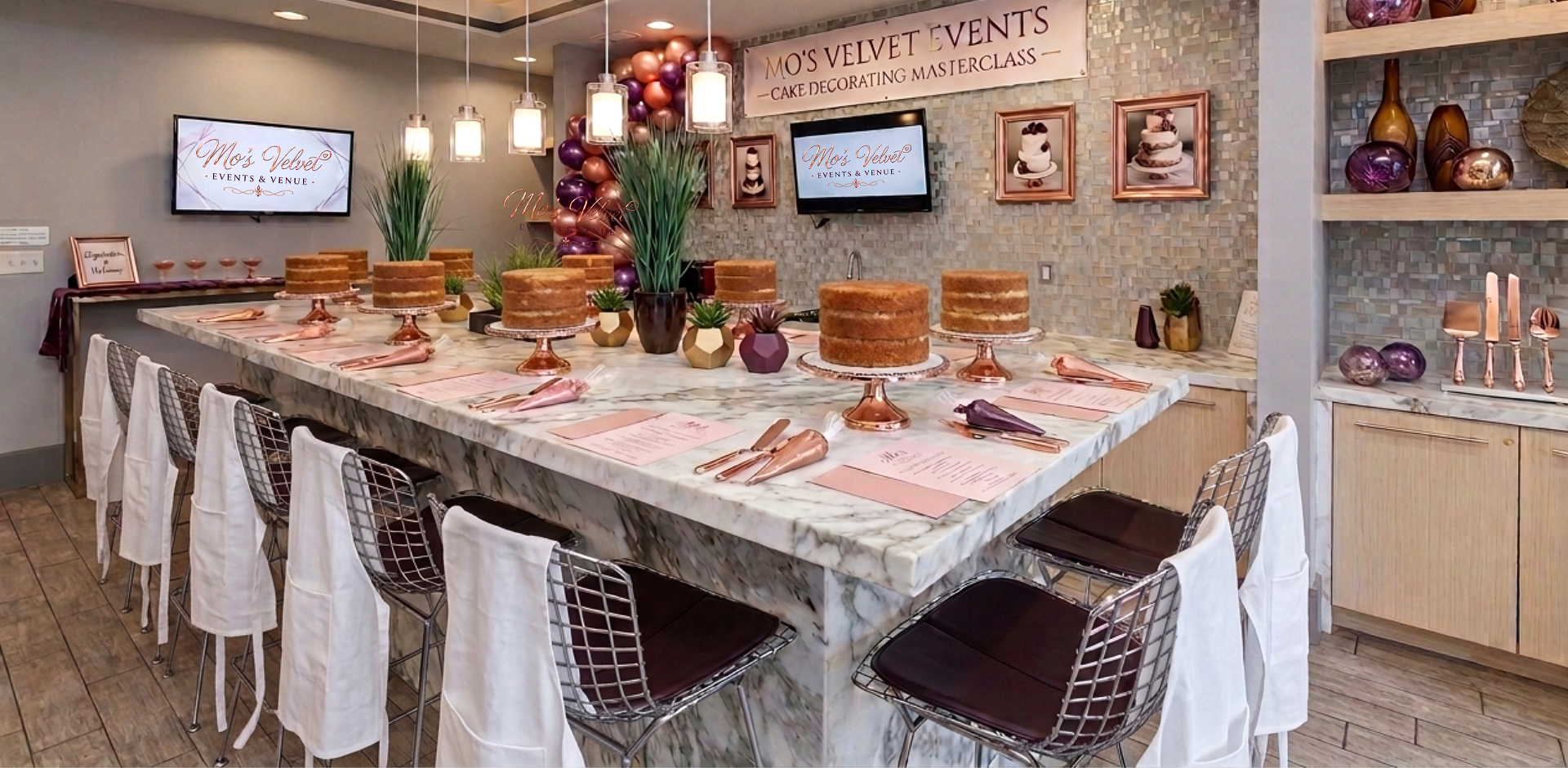 A decorated cake table at an event venue called Mo's Velvet Events, with several naked cakes on rose gold cake stands, pink printed menus, pink and purple napkins, with balloons and framed photos of cakes in the background.