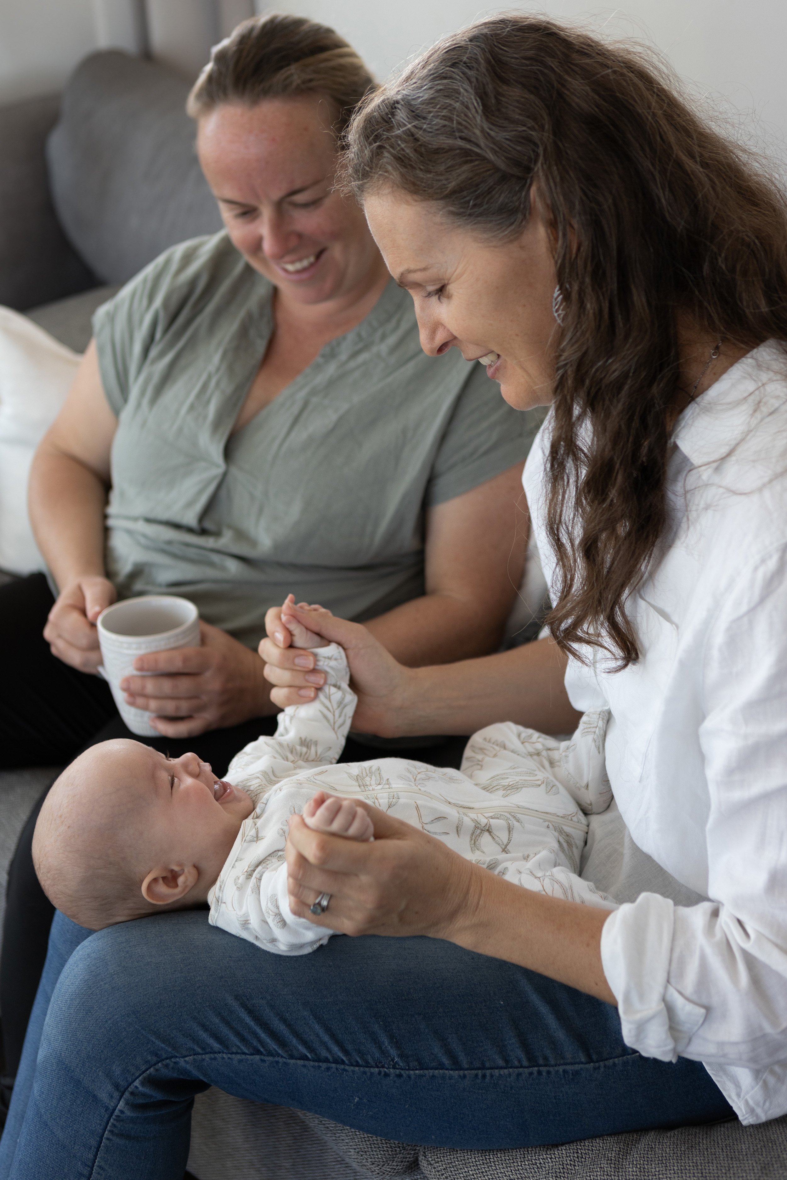 Two women sitting on a gray couch, smiling and looking at a baby lying on a person's lap, who is also smiling and looking up at them. One woman is holding a mug, and the scene is indoors with natural light.
