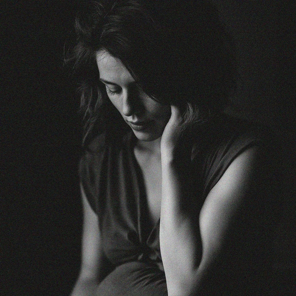 A black and white portrait of a woman with dark hair, her eyes closed, and her hand touching her neck, with soft lighting creating shadows on her face.