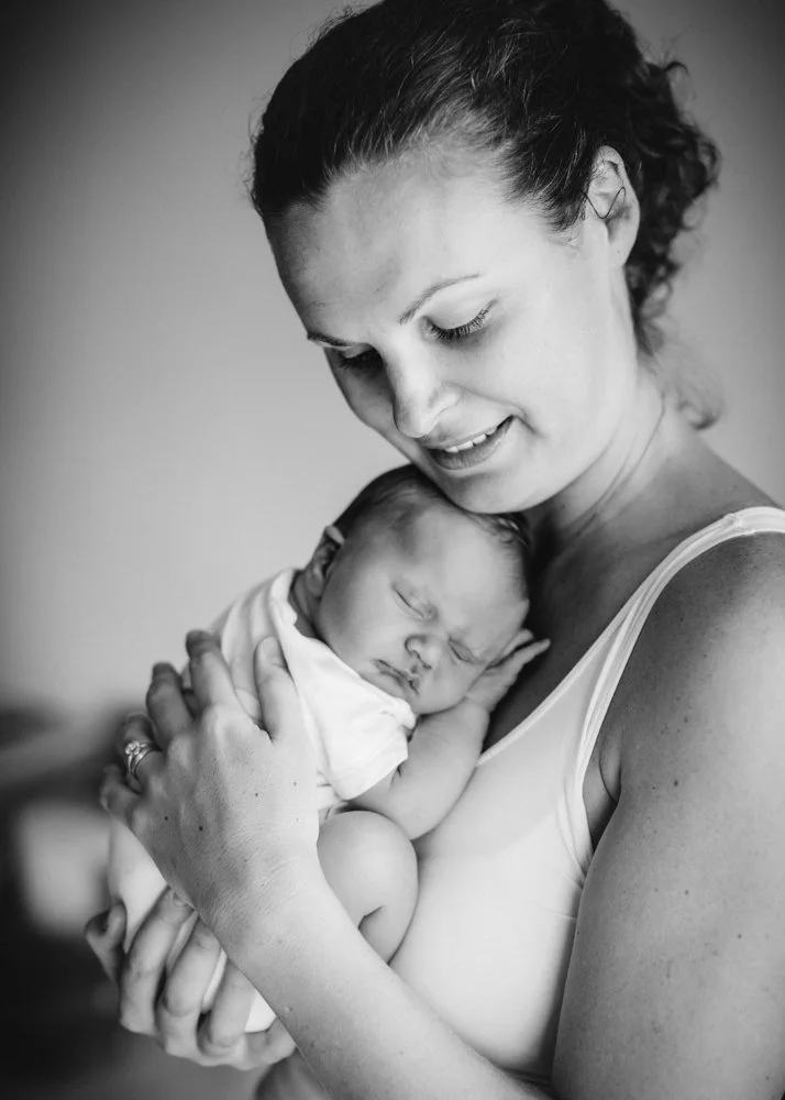A black and white photo of a woman holding a sleeping newborn baby close to her chest, smiling gently.