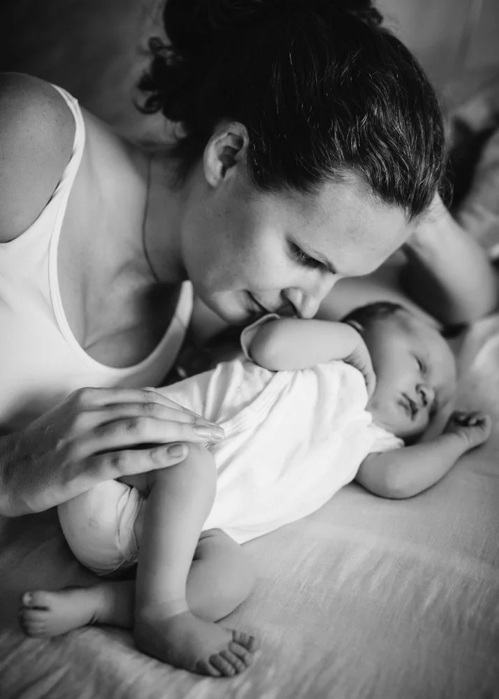 A woman gently kisses a sleeping young child lying on a bed, with another child partially visible in the background.