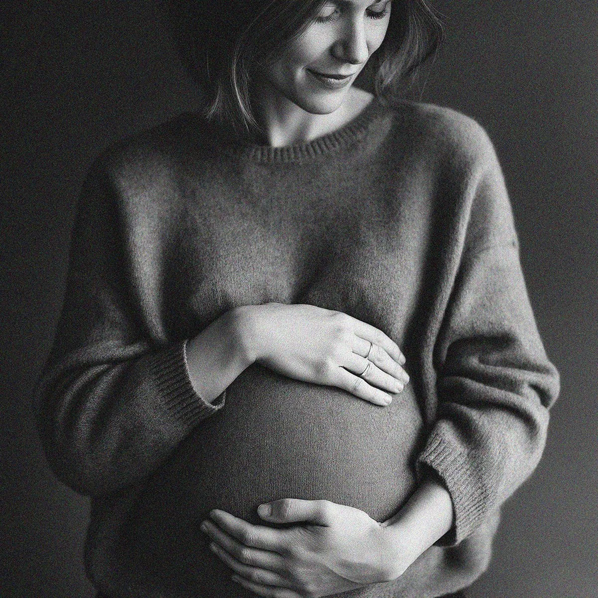 A pregnant woman gently holding her belly with both hands, wearing a cozy sweater, smiling softly, in black and white.