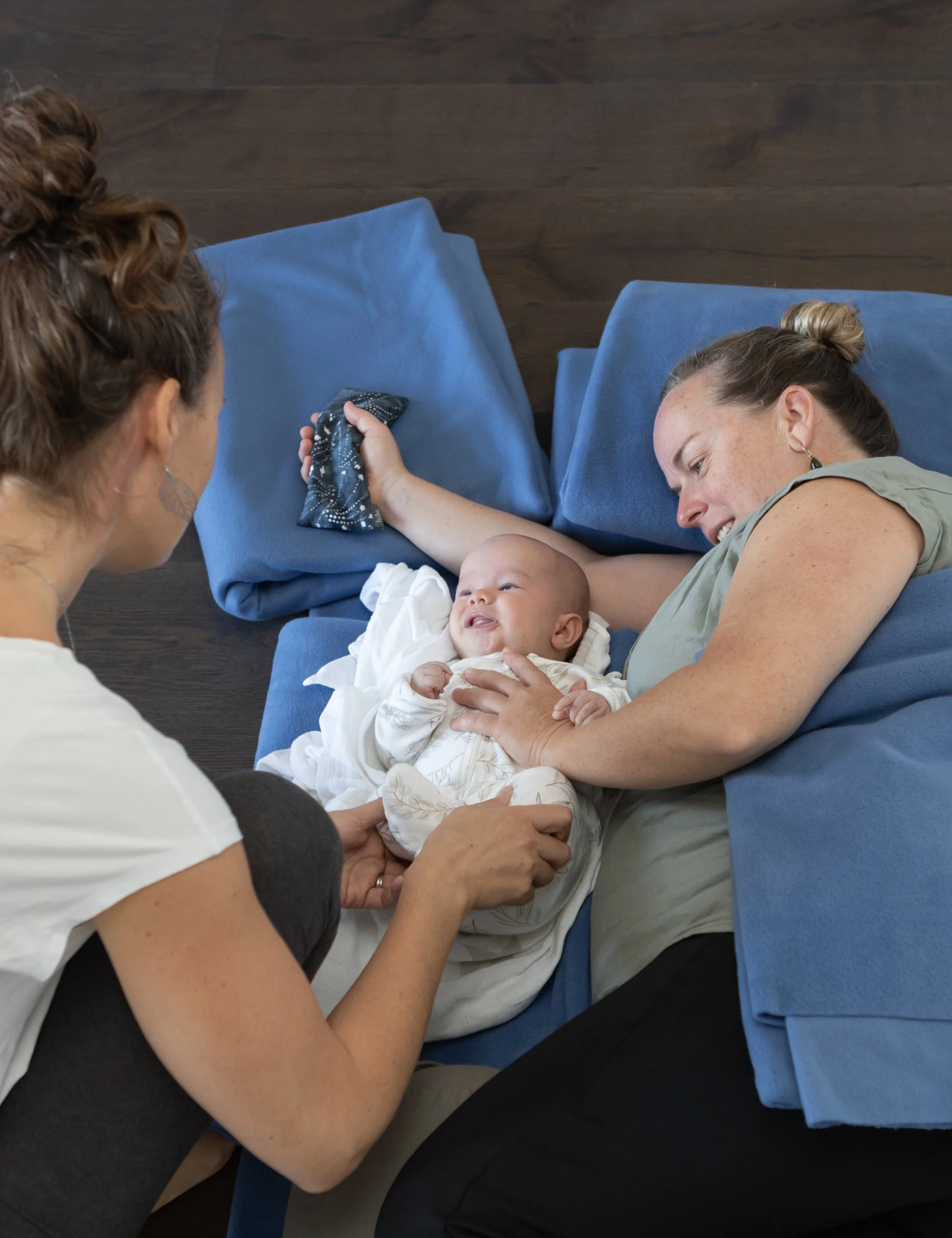 A woman with a bun hairstyle lying on a blue couch, smiling and holding a baby who is also laying on the couch, with another woman in the foreground reaching towards the baby.