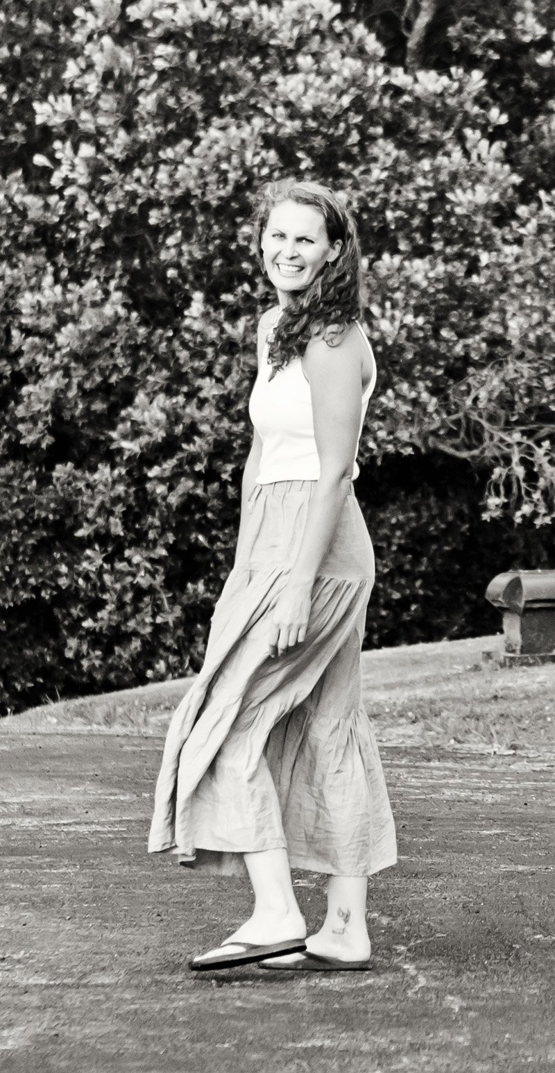 A woman with curly hair smiling and winking, wearing a sleeveless top and a long skirt, standing outdoors in front of bushes.