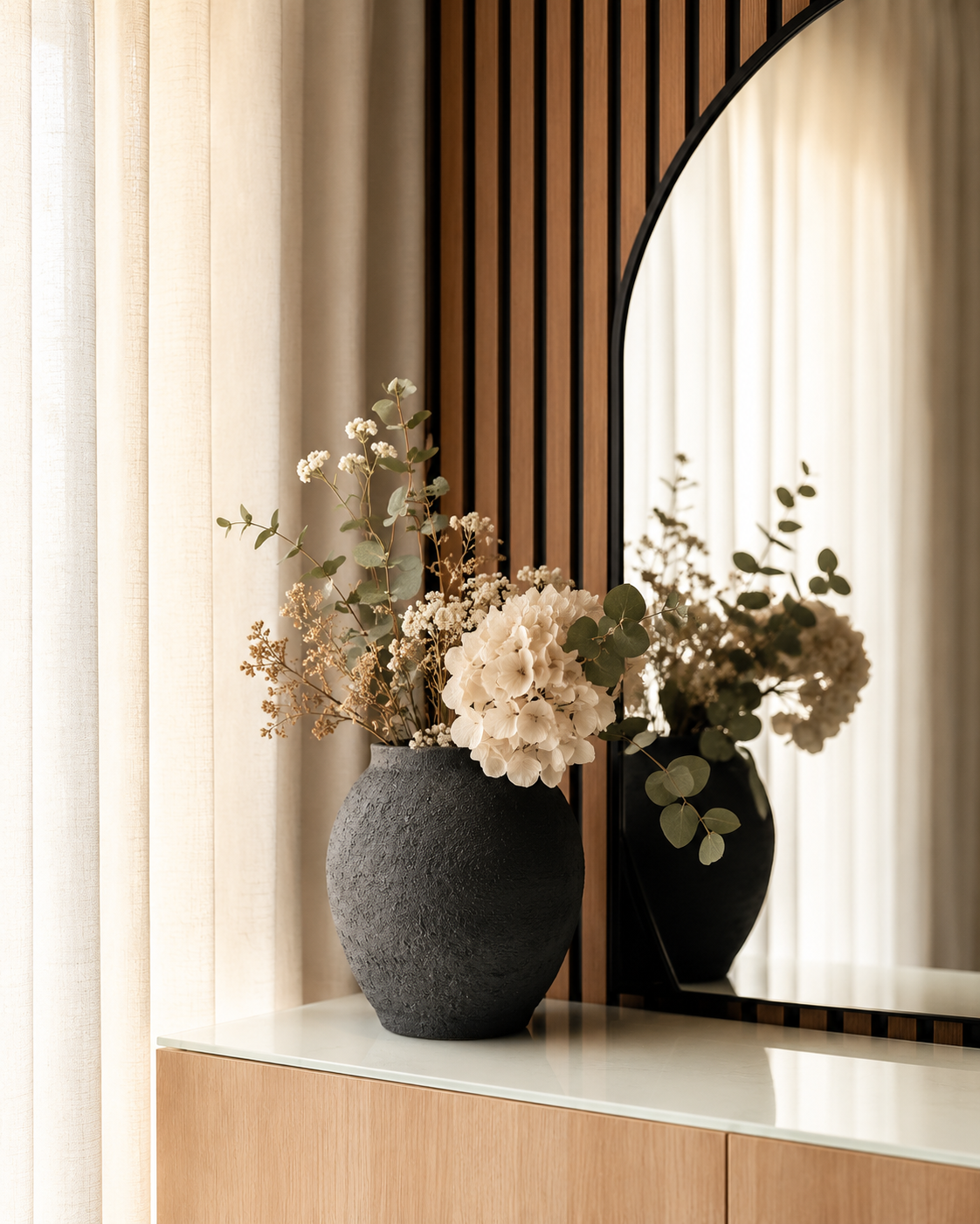 A black textured vase with white hydrangeas and eucalyptus leaves on a white and wood side table, reflected in a large mirror with wooden slats and beige curtains in the background.