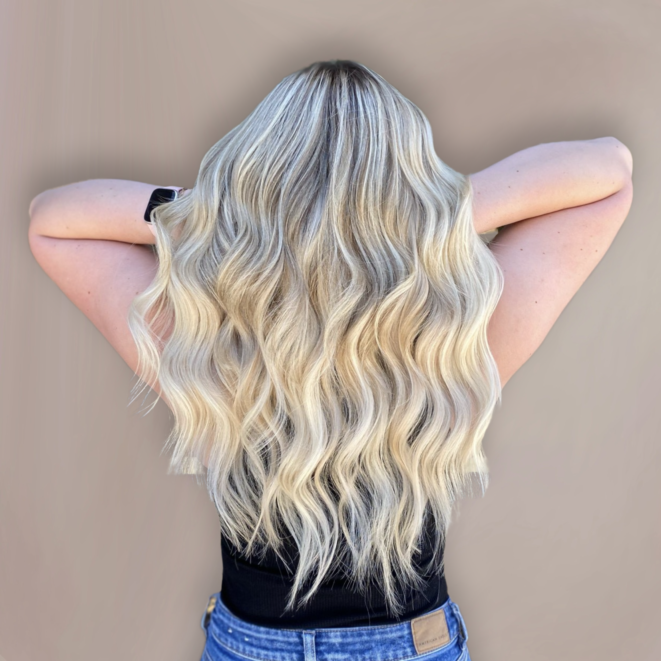 A person with long, wavy blonde hair taking a selfie from the back, wearing a black shirt and blue jeans, with a plain beige wall in the background.