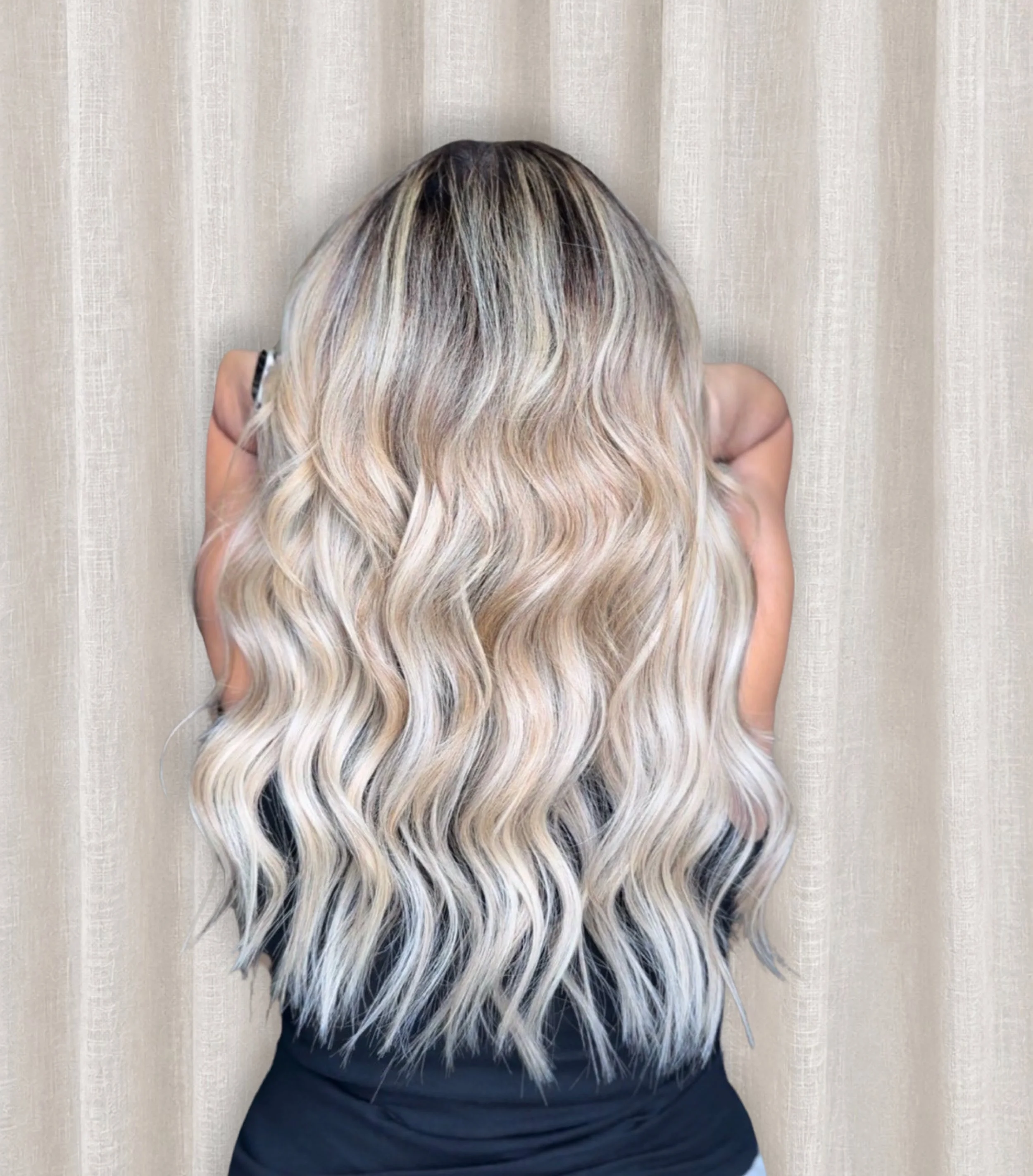 A woman with long, wavy hair featuring dark roots and blonde tips, seen from behind, against a beige curtain background.