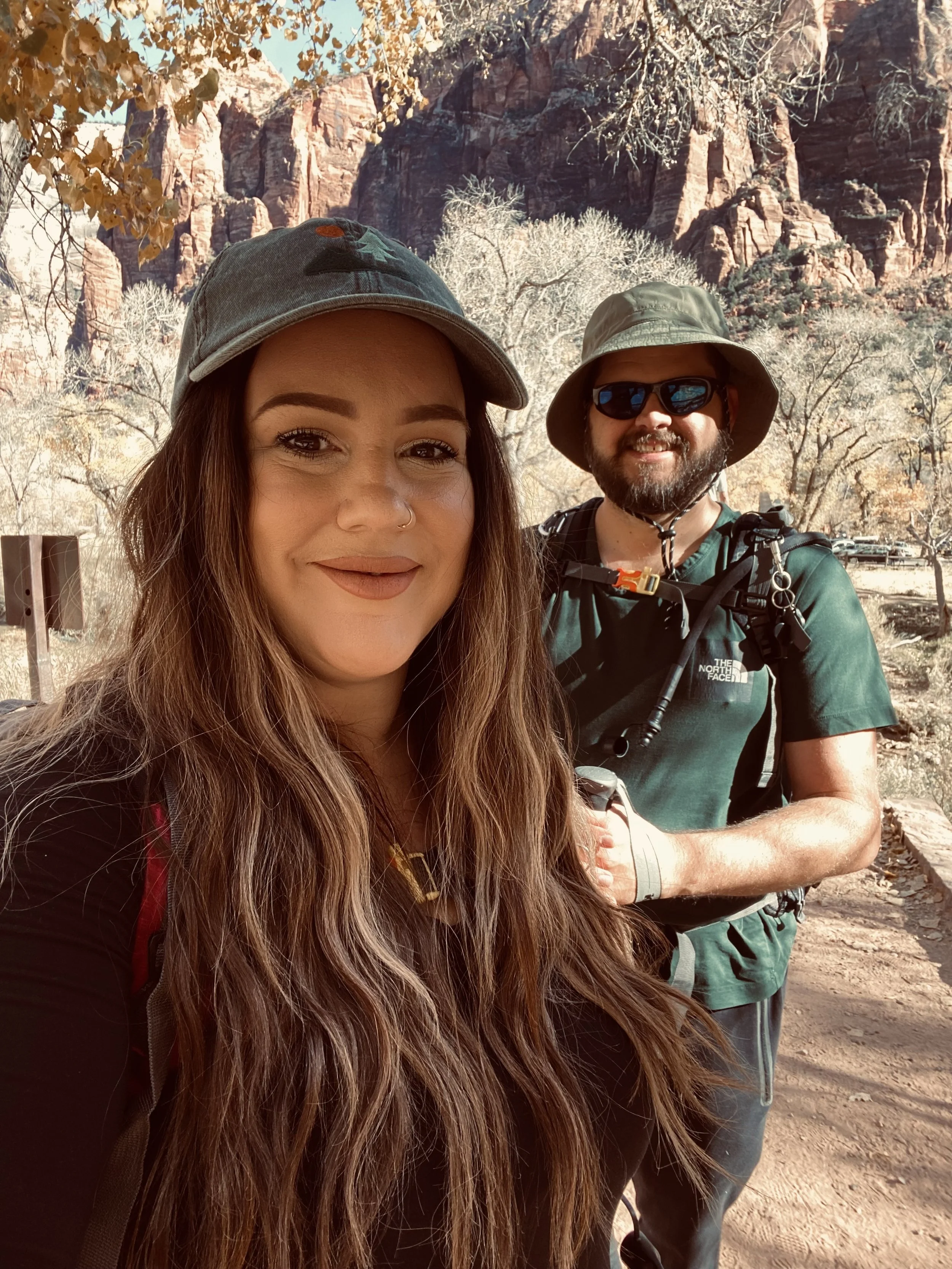 A woman and a man smiling outdoors, with a rocky red canyon and leafless trees in the background. The woman is in the foreground, wearing a gray cap and long hair. The man is wearing sunglasses, a green hat, and a green shirt with a backpack.