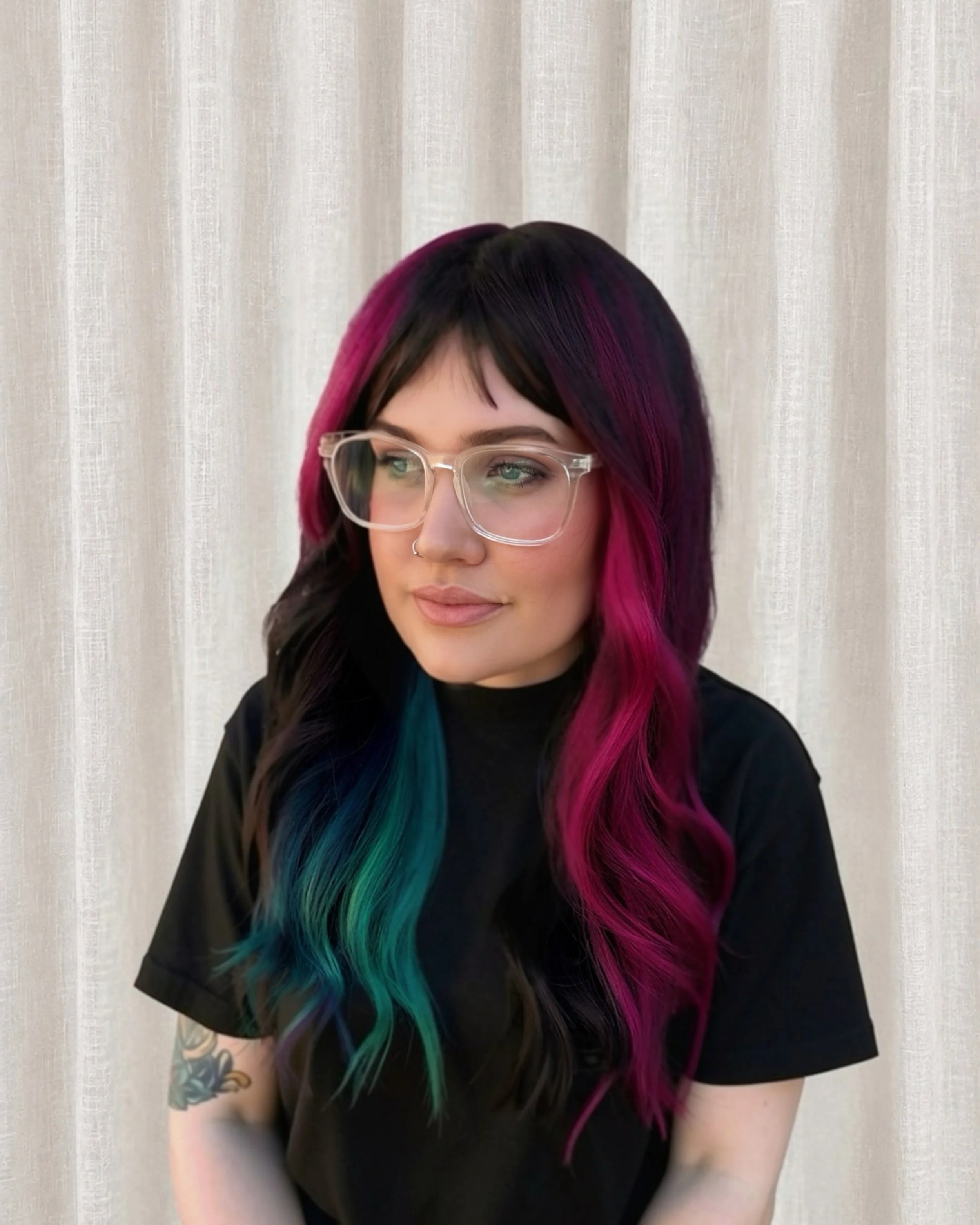 A woman with black hair that fades into bright pink, purple, and teal at the ends, wearing glasses and a black shirt, sitting in front of a light-colored curtain.