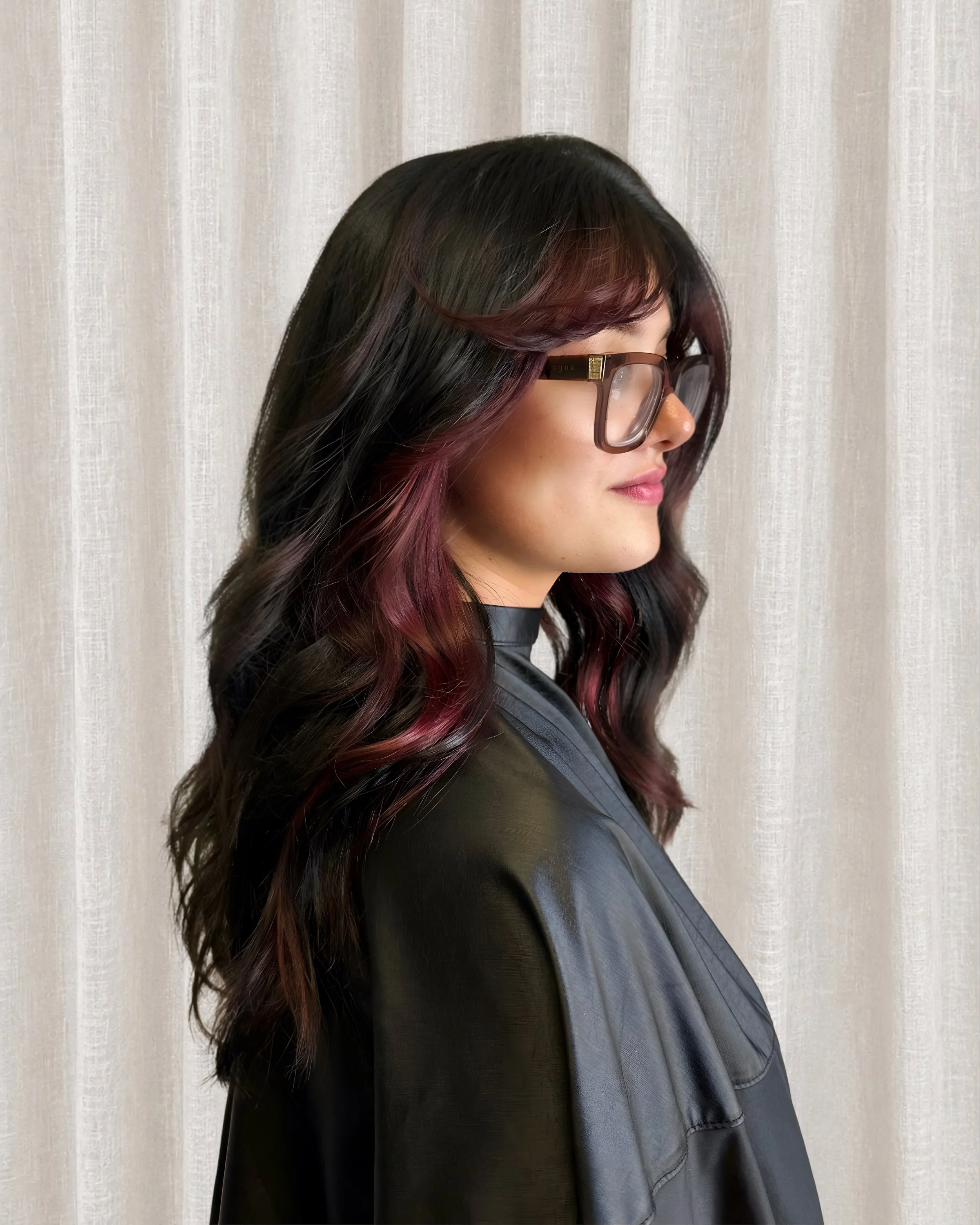 Woman with long, wavy black and burgundy hair wearing large, rectangular glasses and a black leather jacket, standing in front of a beige curtains background.