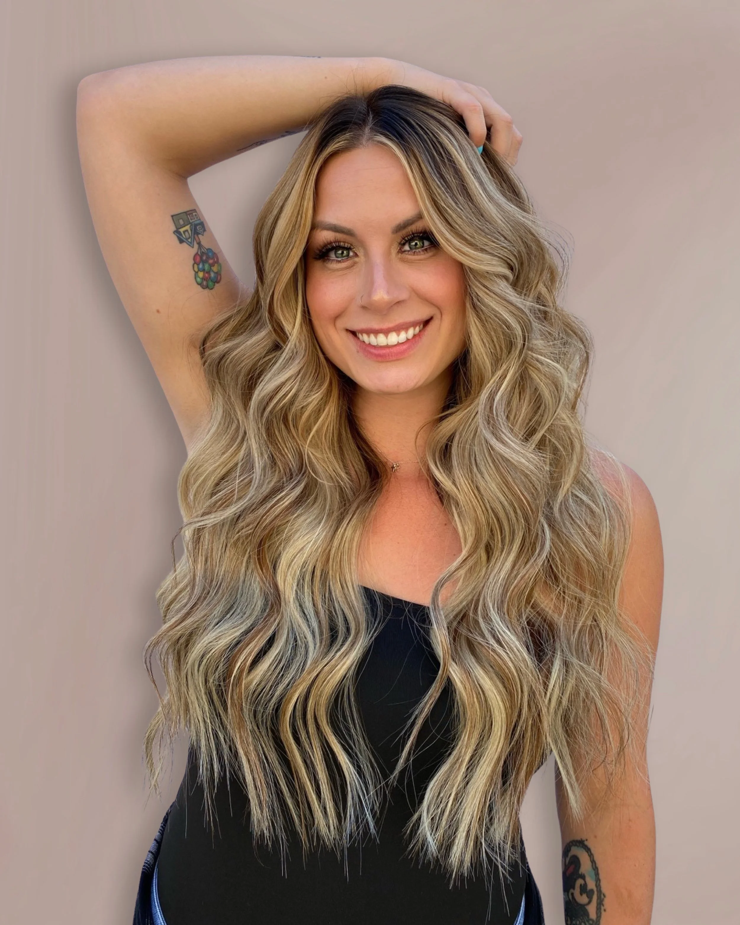 A woman with long, wavy blonde hair, smiling, with her right hand resting on her head. She has tattoos on both her right arm and left forearm, and is wearing a black top and a small necklace. The background is neutral and plain.