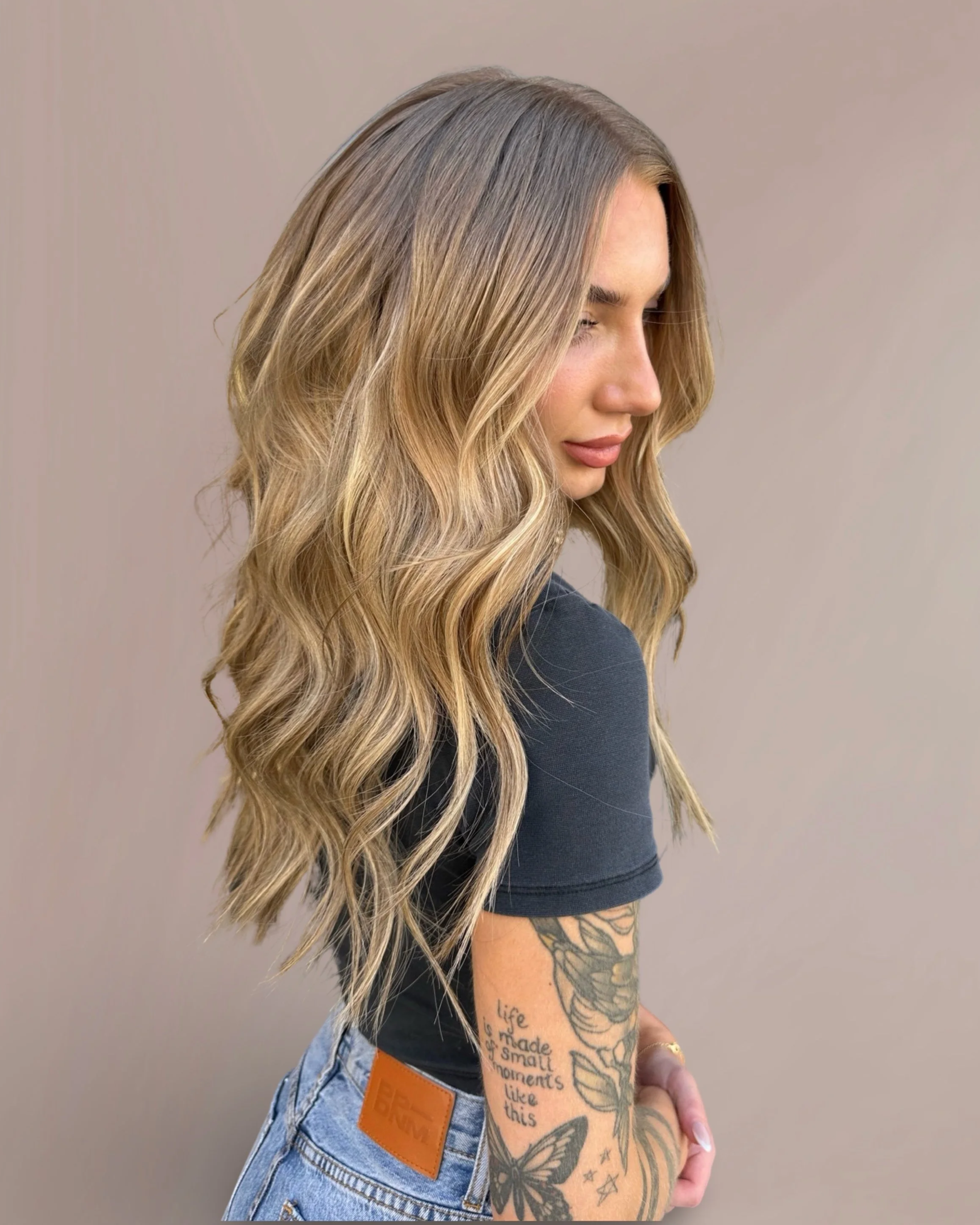 A woman with long blonde wavy hair looking down, wearing a black shirt with tattoos on her arm, and light blue jeans with a brown leather patch on the waistband, set against a plain beige background.