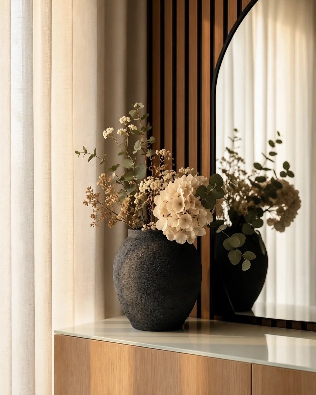 A black textured vase with dried white flowers and green foliage on a wooden surface, with a large mirror behind reflecting the flowers and a cream-colored curtain.