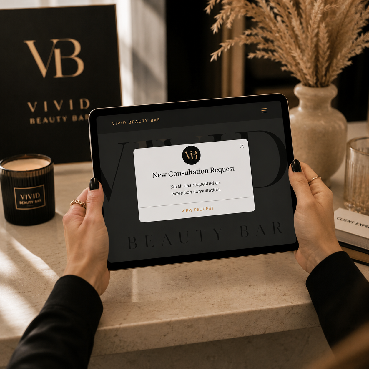 A person holding a tablet with a notification for a new consultation request at Vivid Beauty Bar. The background shows a black Vivid Beauty Bar bag, a candle, a vase with dried flowers, and an informational booklet on a countertop.