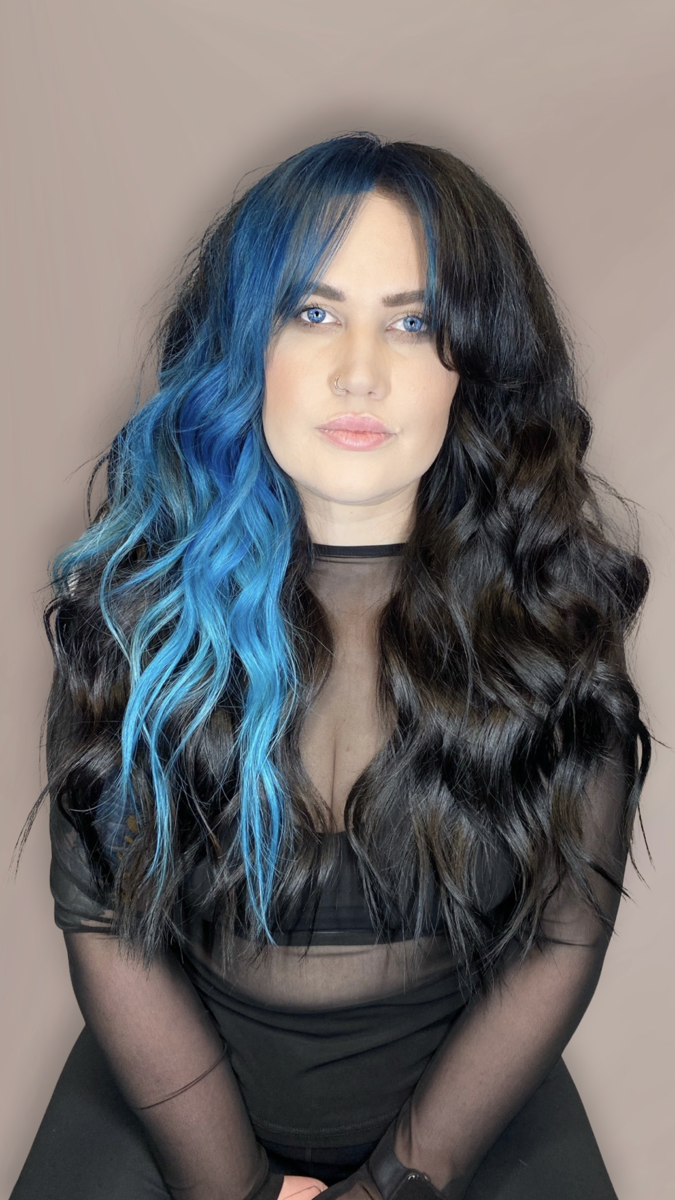A woman with long wavy black hair with a streak of bright blue, wearing makeup, a nose ring, and black sheer top, sitting against a plain background.