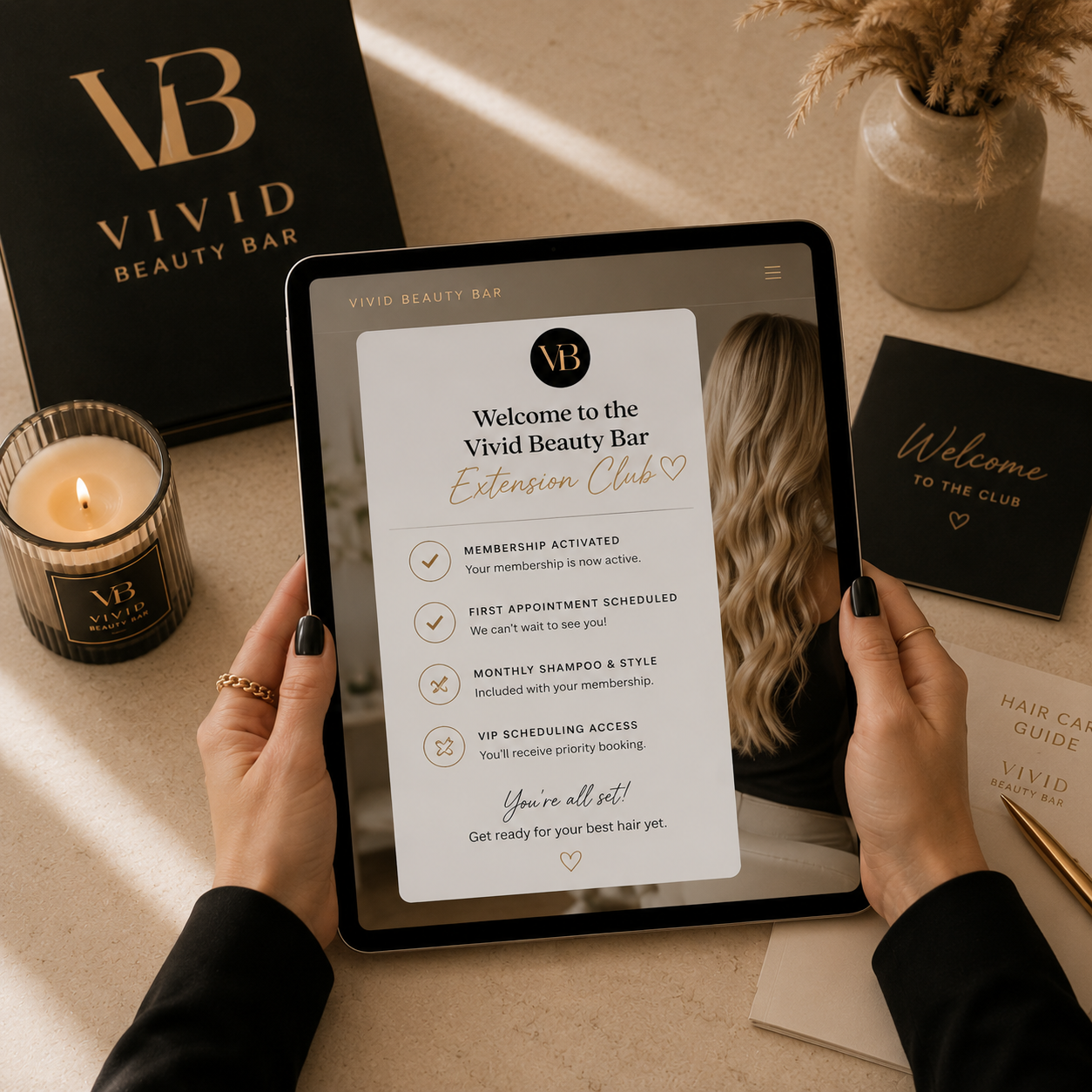 Person holding a tablet displaying the Vivid Beauty Bar Extension Club membership details. Around the tablet on the beige surface are a lit candle with a black and gold label, a black and gold welcome card, a hair care guide, and a beige vase with dried pampas grass.