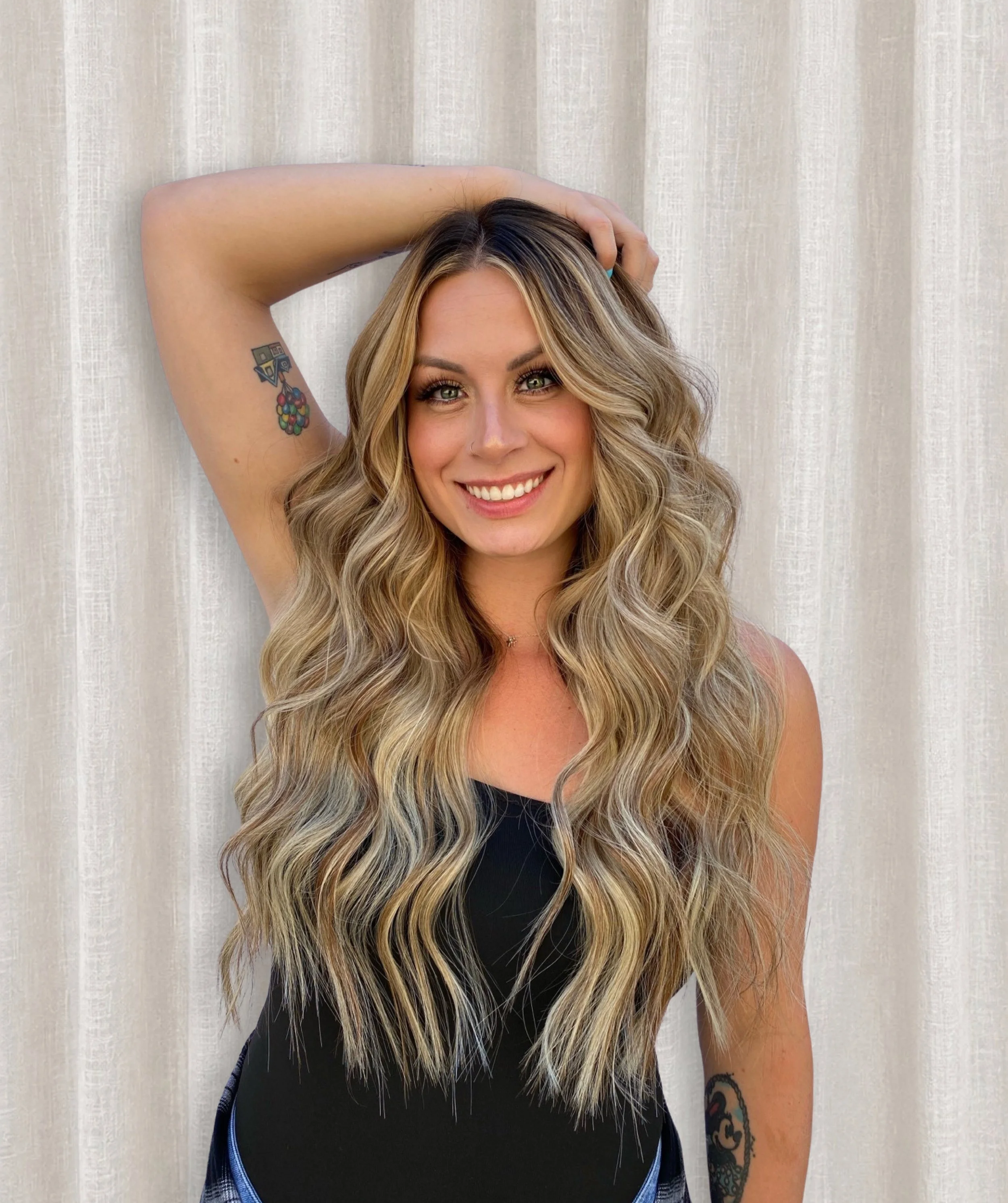 A woman with long, wavy blonde hair, smiling, with a light-colored curtain background, wearing a black top and showing tattoos on her left arm.