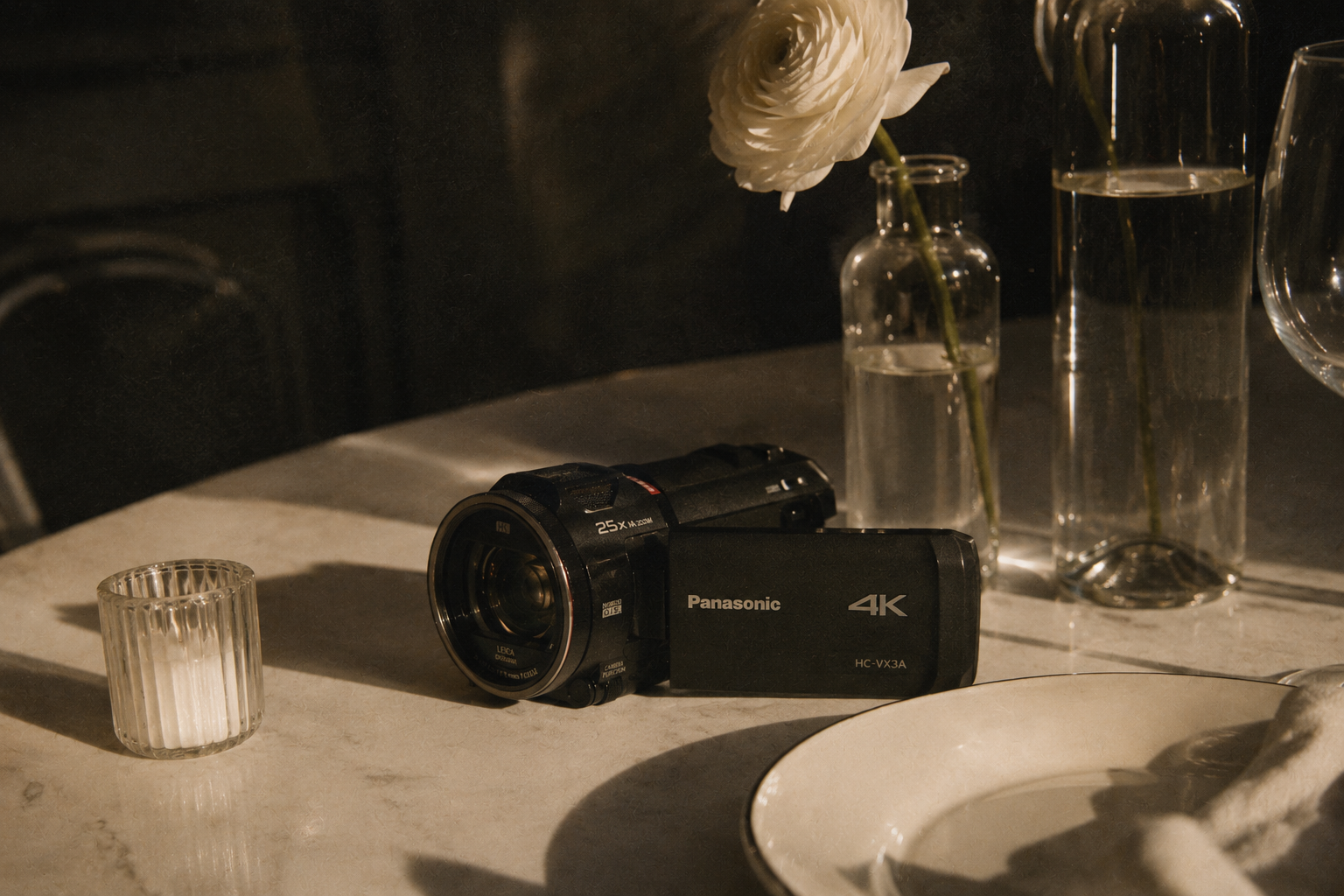 A black Panasonic 4K camcorder placed on a white marble table, next to a small ribbed glass container, a white flower in a tall clear glass vase, and a larger clear glass vase, with additional glassware in the background.