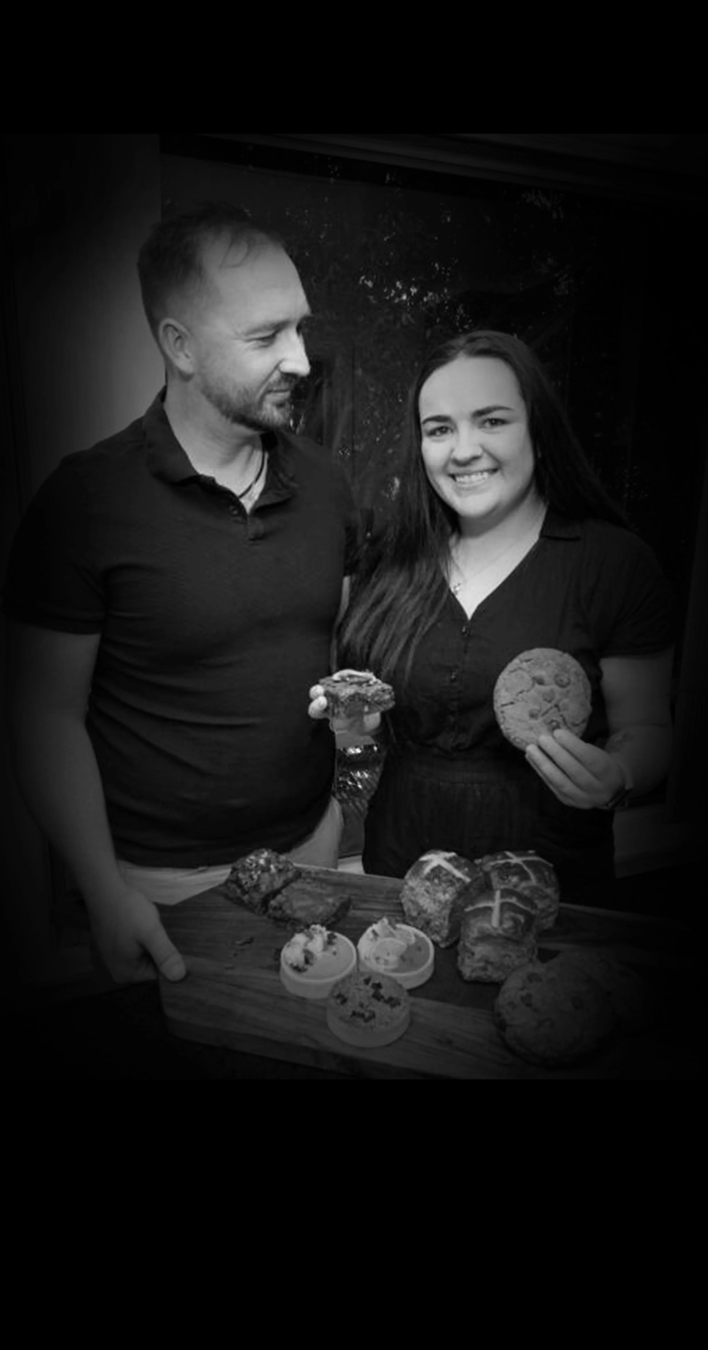 A man and woman standing behind a table filled with cupcakes, cookies, and baked goods, with the woman holding a cookie and the man holding a piece of cake, smiling and looking at each other.